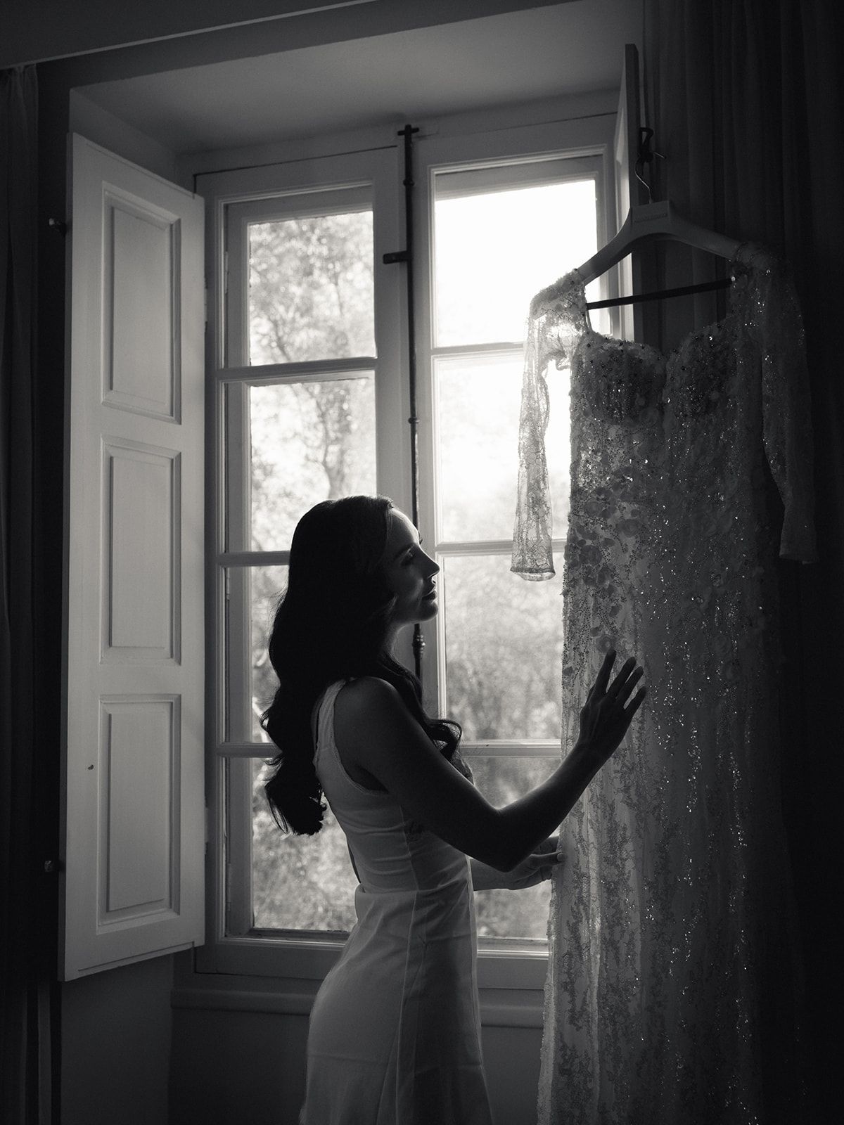 Bride silhouetted at the window beside her hanging gown, black and white