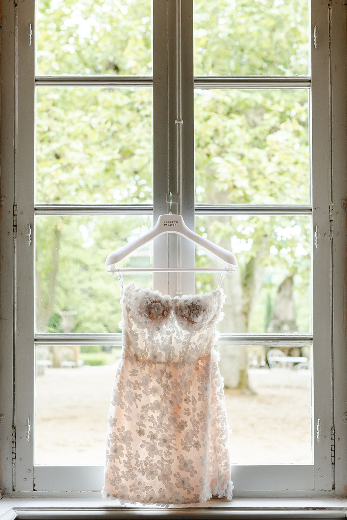 Short blush dress with a floral bodice hanging in a tall window at Château de Pradines