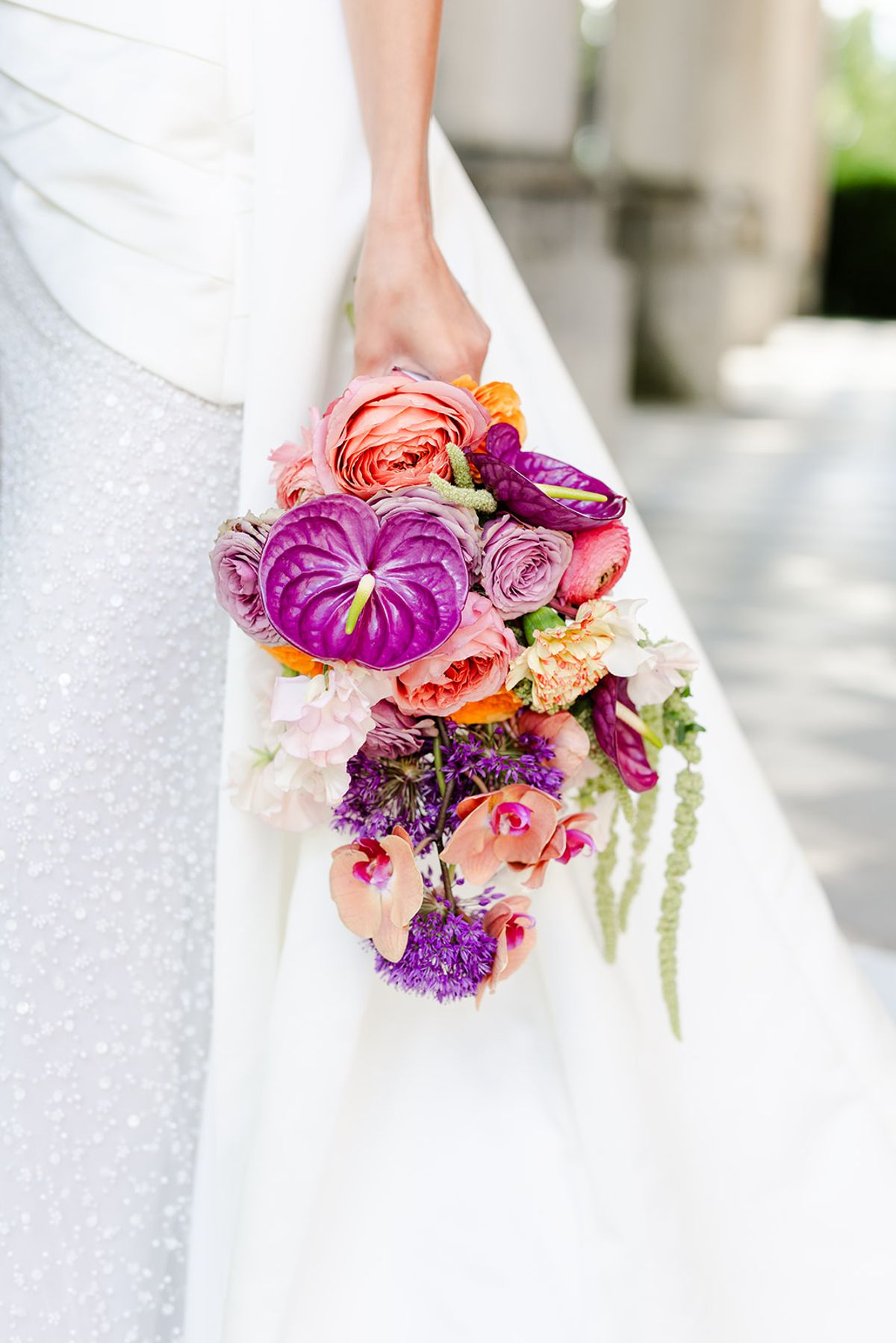 Bridal bouquet of magenta anthurium, coral roses and purple flowers against a white gown