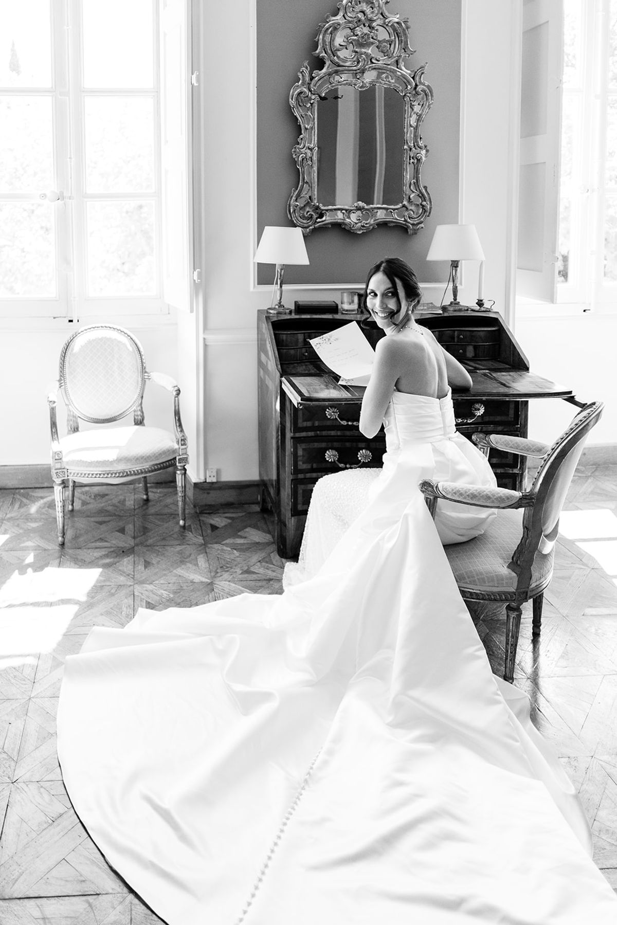 Bride seated at a writing bureau with her train spread across the floor, black and white