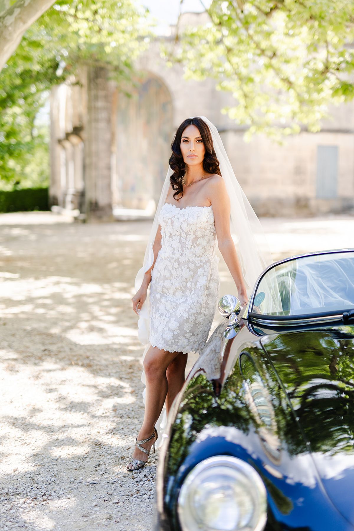 Bride in a short blush floral dress and long veil beside a dark blue vintage car