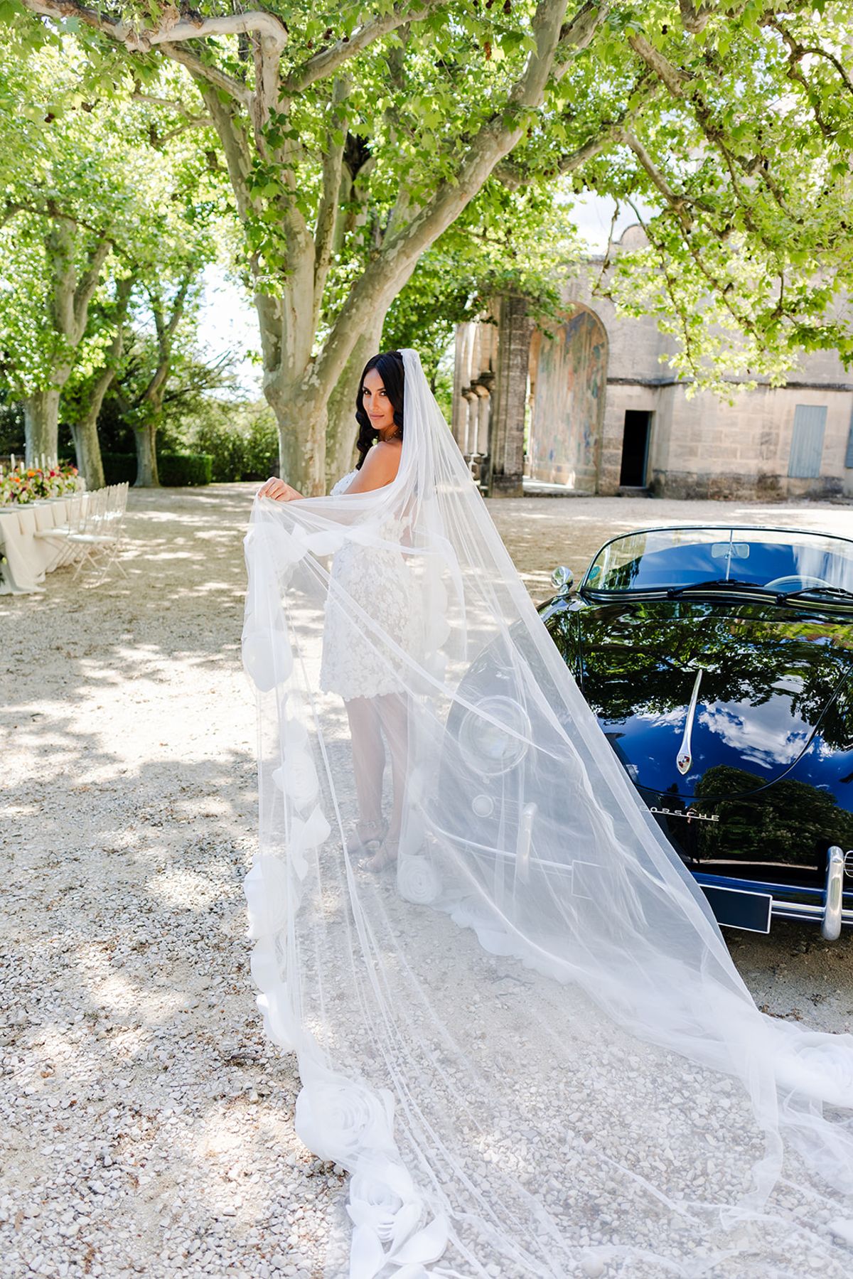 Bride holding out a cathedral-length veil beside a vintage car under plane trees