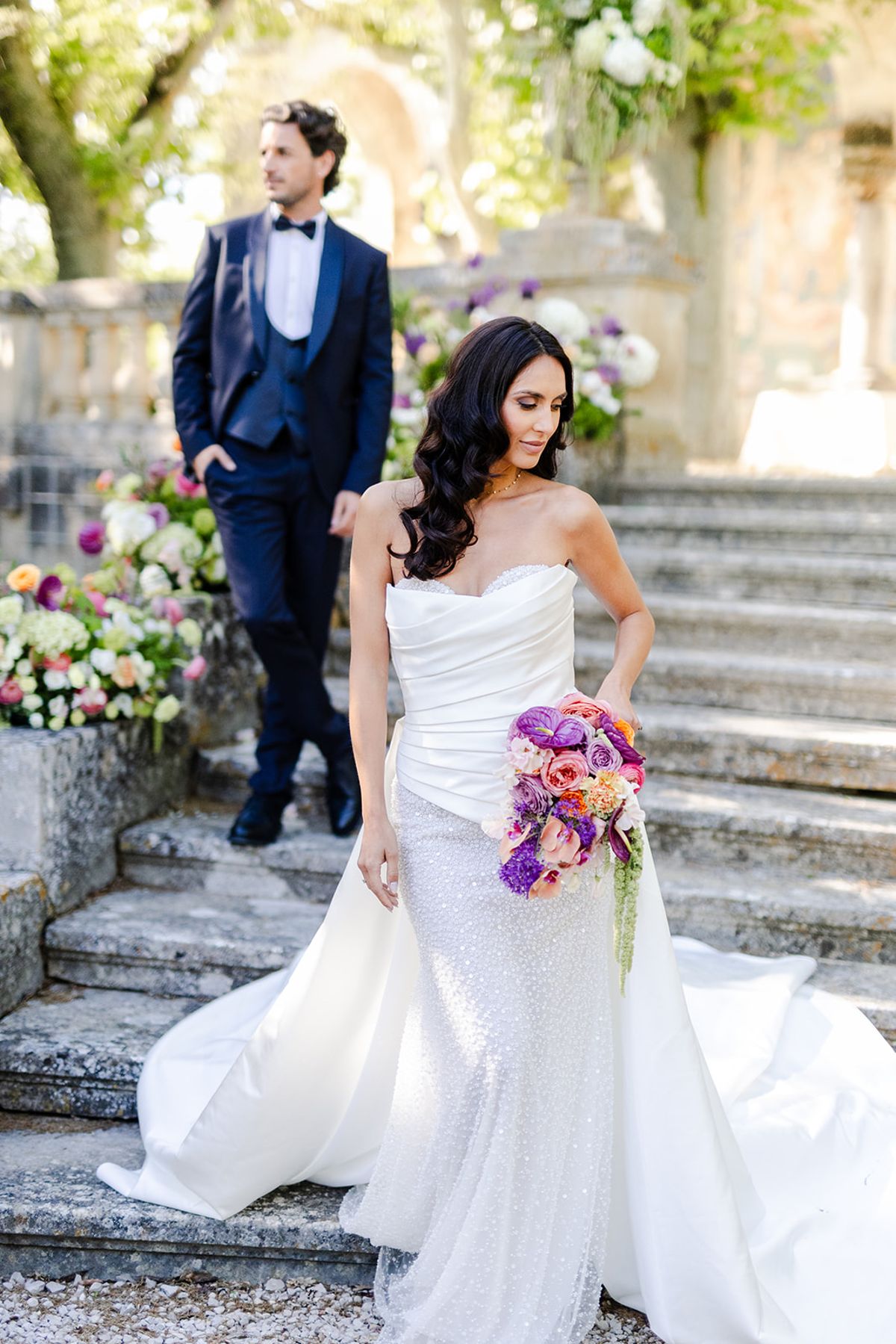Bride in a beaded gown with a satin overskirt on the staircase, the groom standing behind her