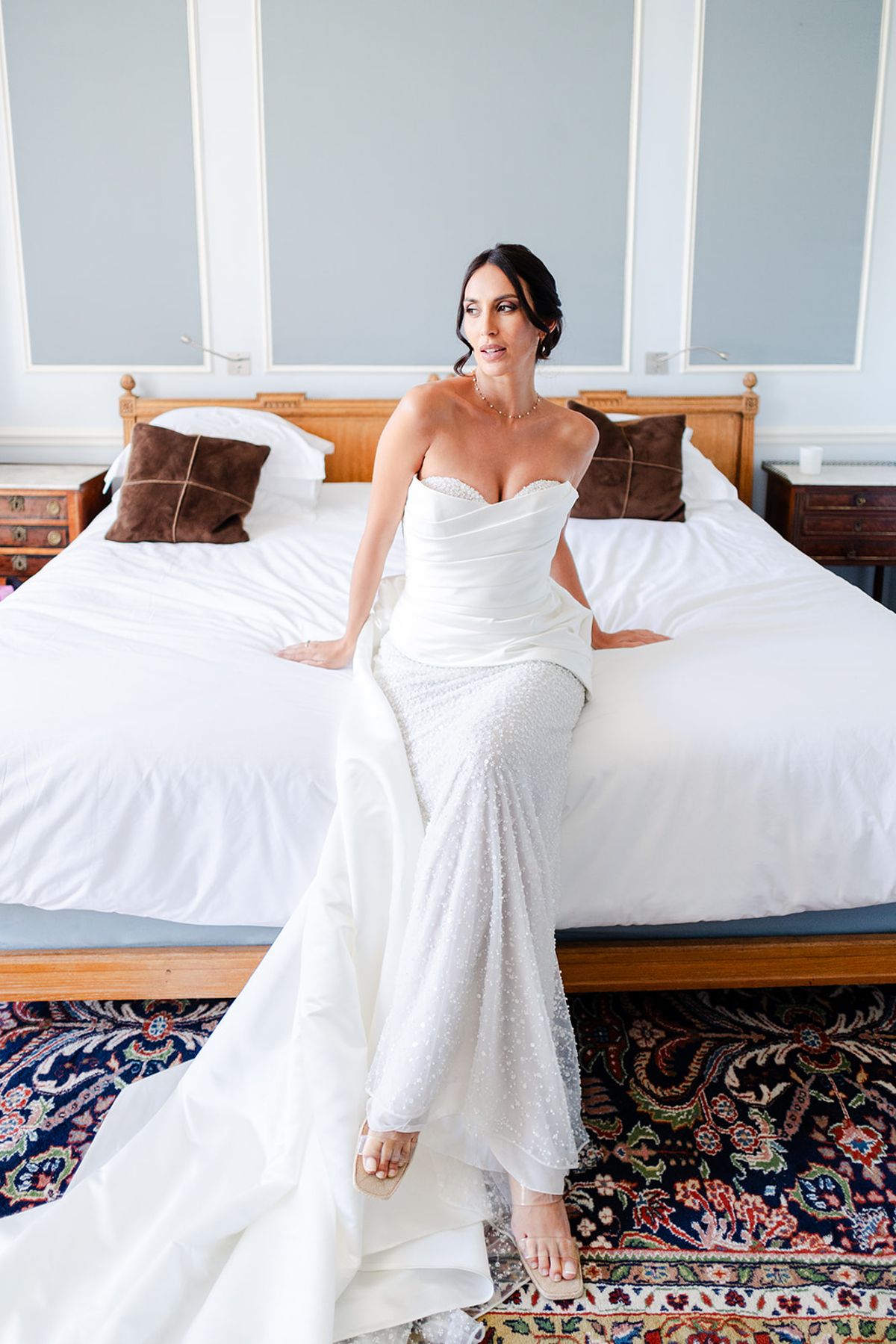 Bride in a fitted white gown sitting on the edge of a bed in a château bedroom