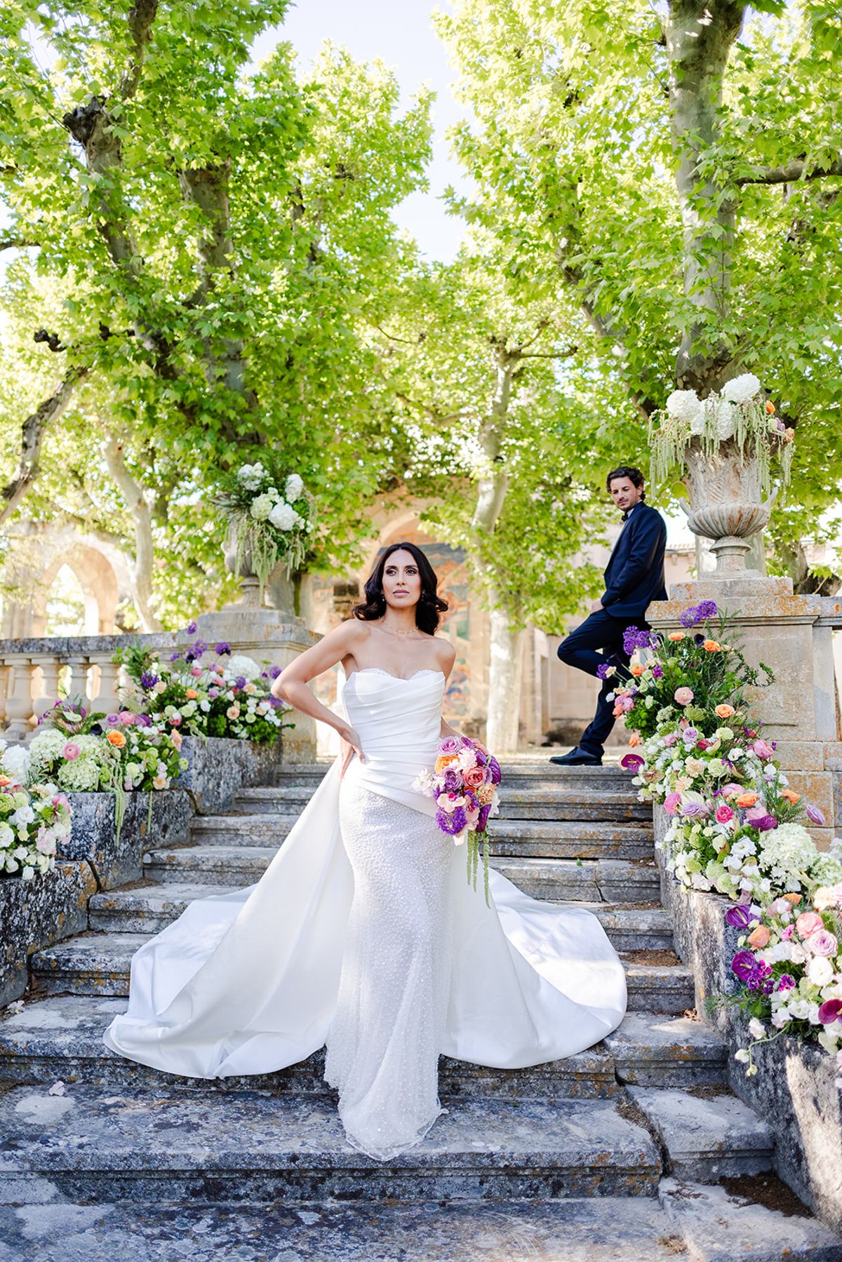 Bride on the garden staircase with a magenta bouquet, the groom leaning on the balustrade behind