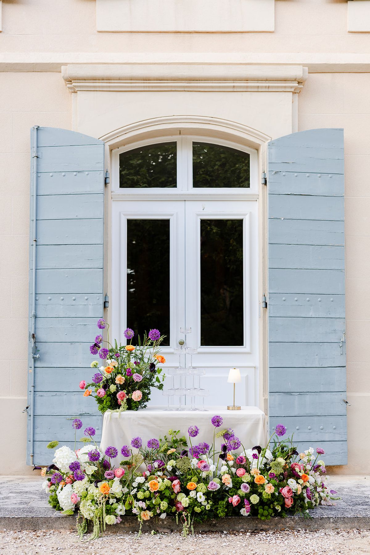 Champagne coupe tower on a flower-banked table in front of blue-grey shutters and French doors