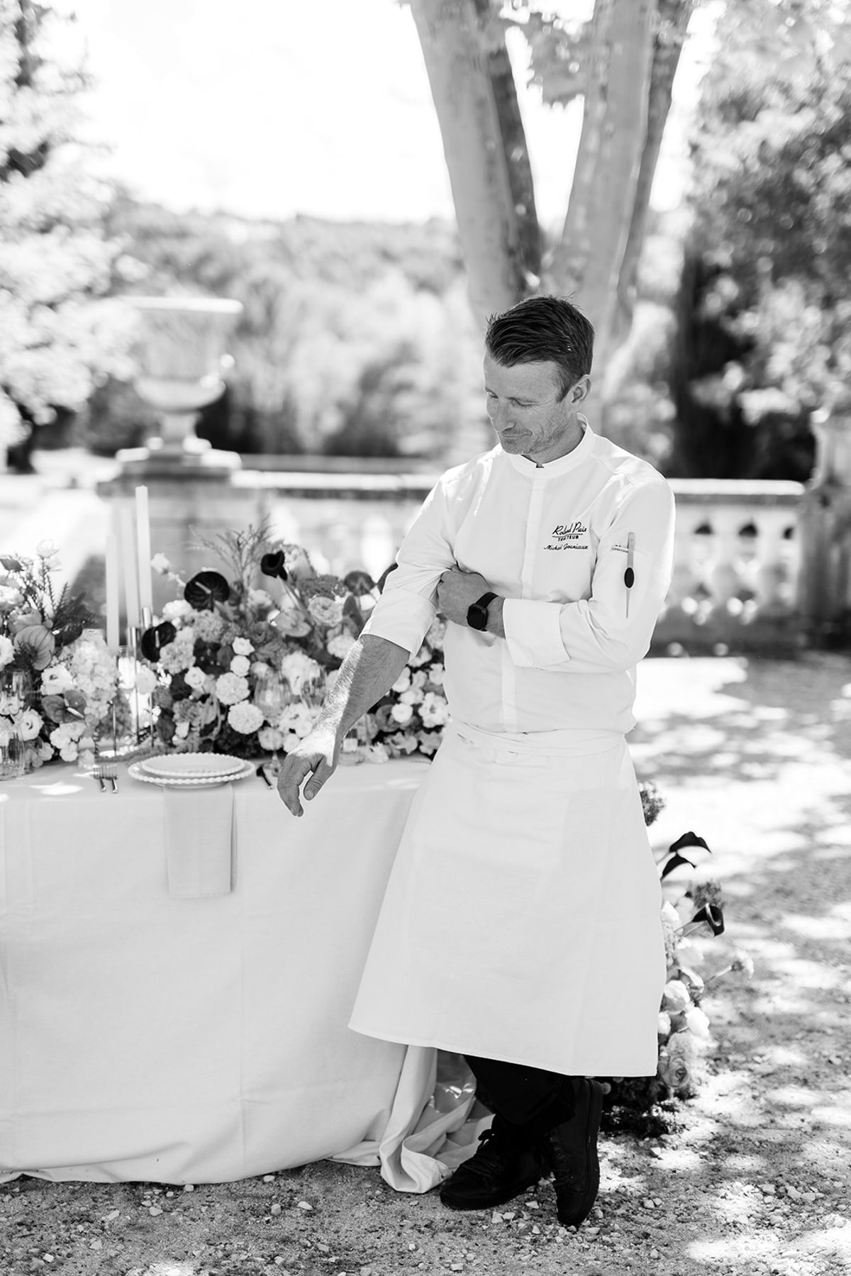 Chef in whites standing at the dinner table beside the flowers, black and white