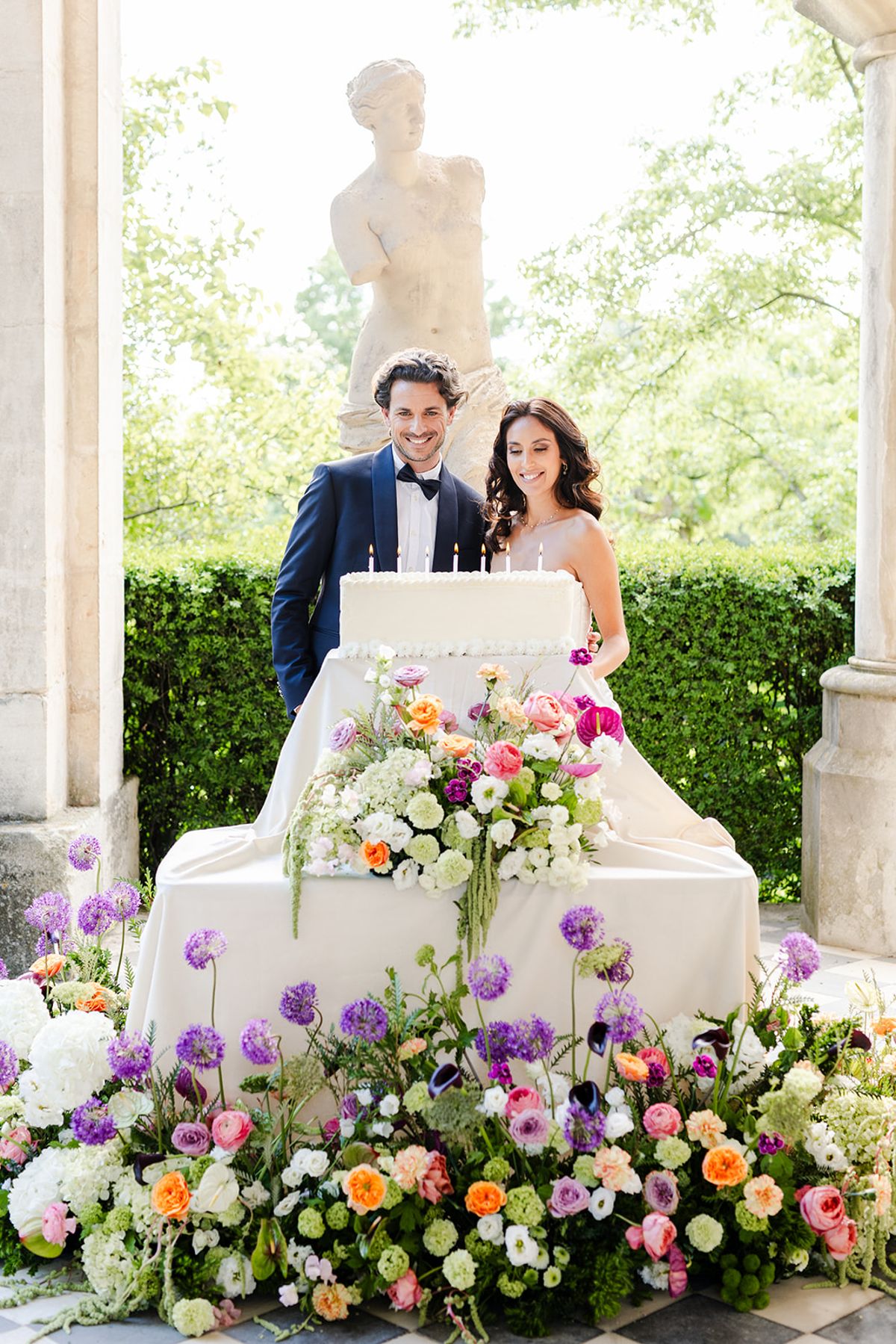 Couple behind a white cake with candles on a draped, flower-banked plinth, a classical statue behind them