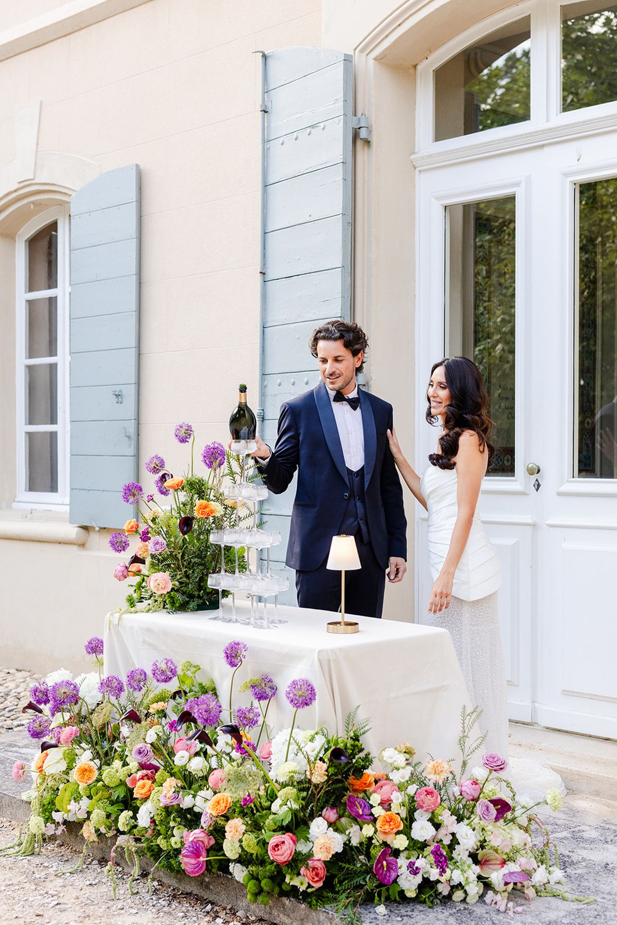 Groom holding a champagne bottle beside a coupe tower and the bride at the French doors