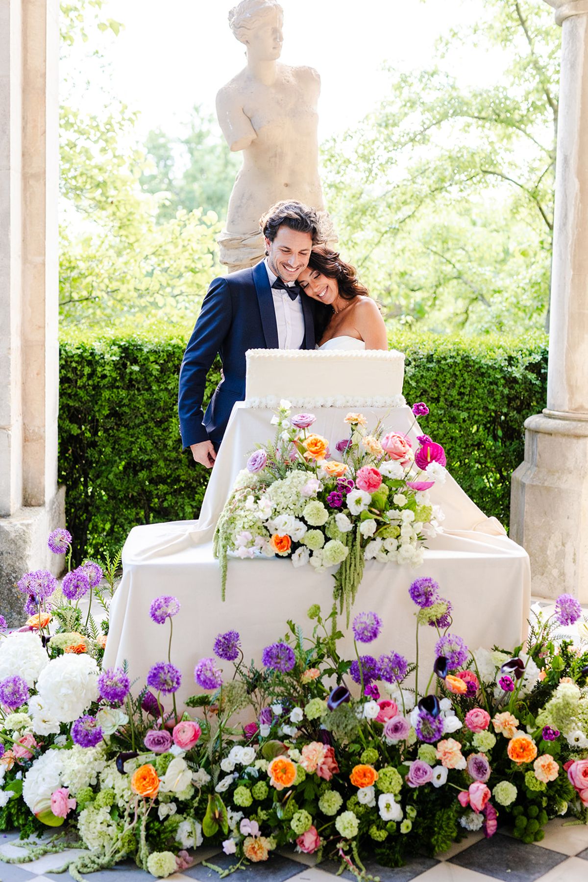 Couple embracing behind the cake with purple allium and roses at its base