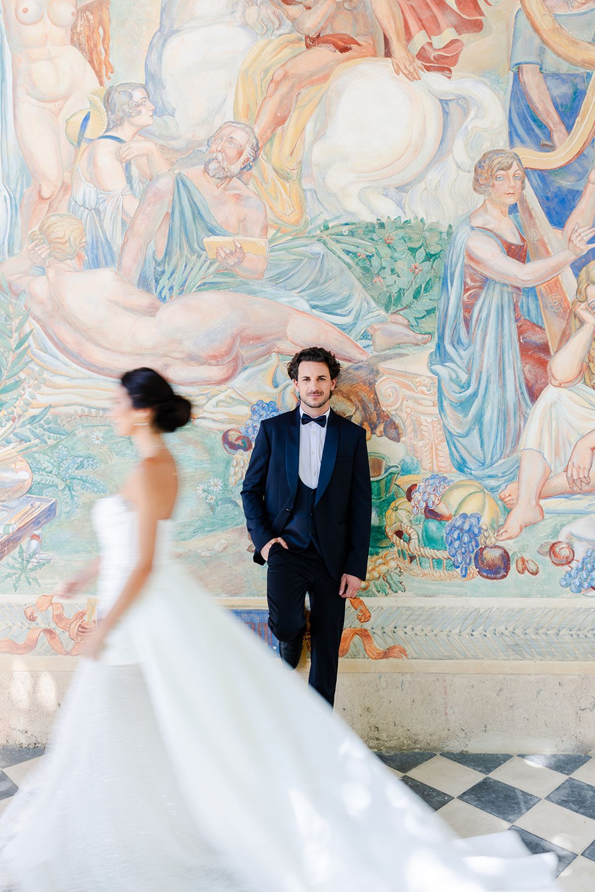 Bride walking past the groom in front of a painted fresco of bathers in pale blue and peach