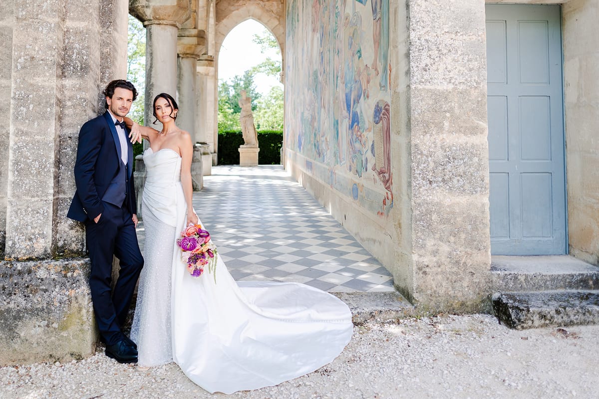 Couple standing in the frescoed loggia above a checkerboard floor, bride holding a magenta bouquet