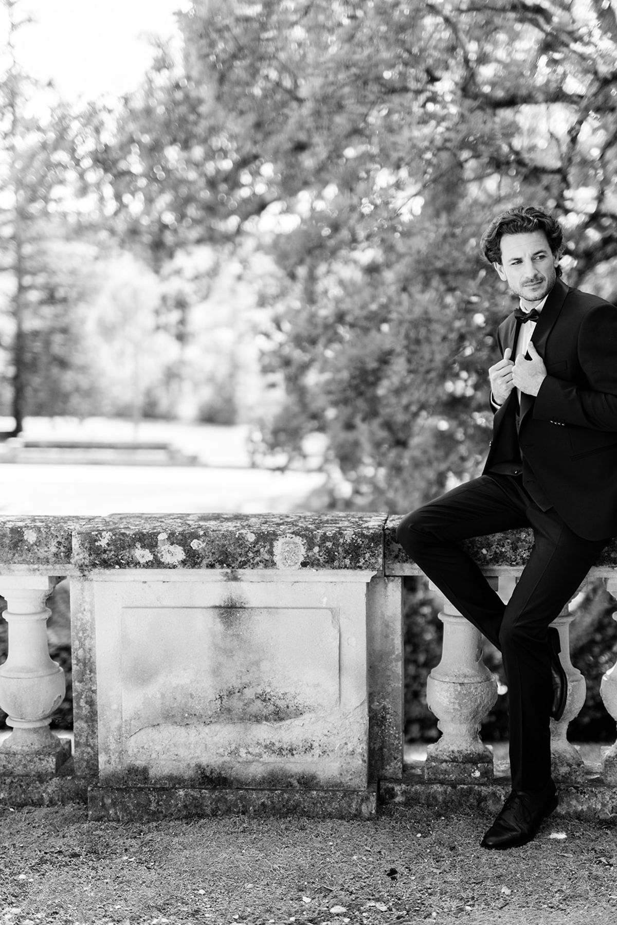 Groom in a tuxedo sitting on a stone balustrade in the gardens, black and white