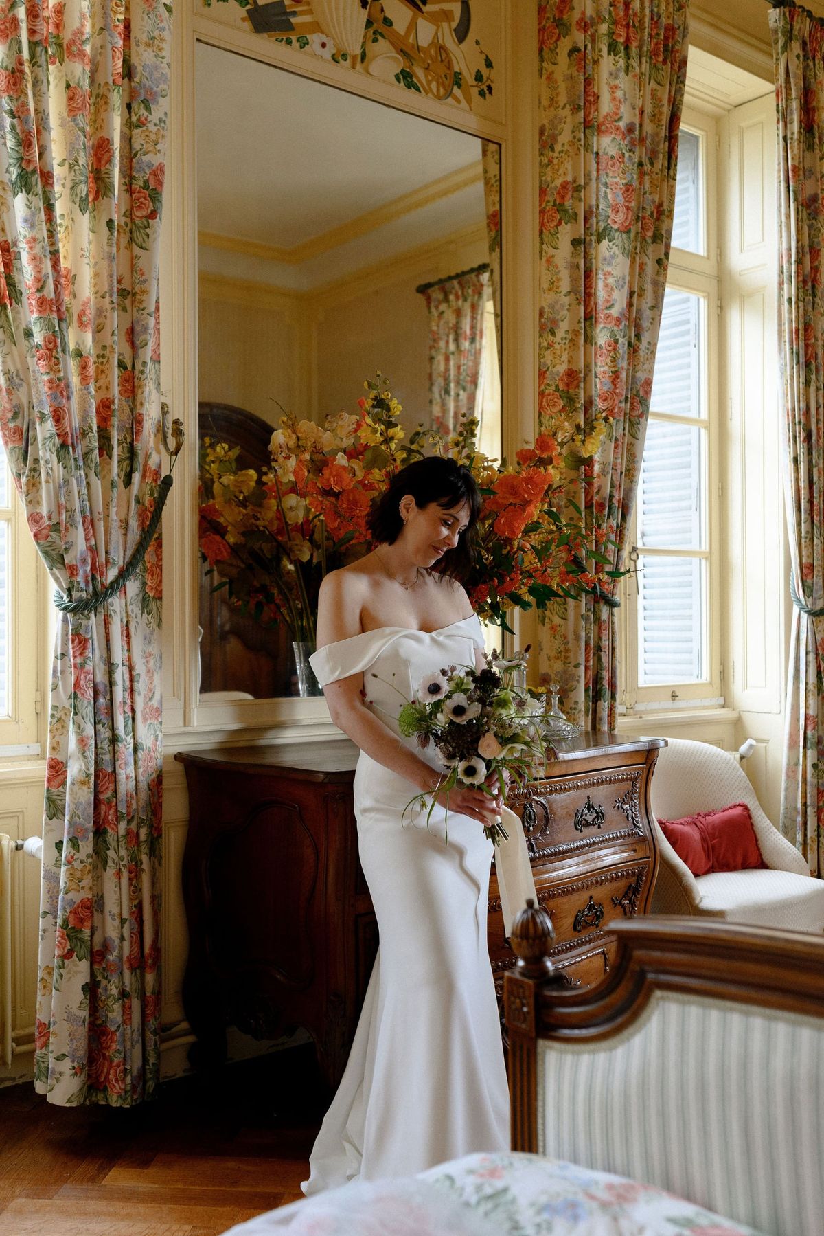 Bride holding anemones beside a large mirror and floral curtains in the Claude suite at Château du Terral