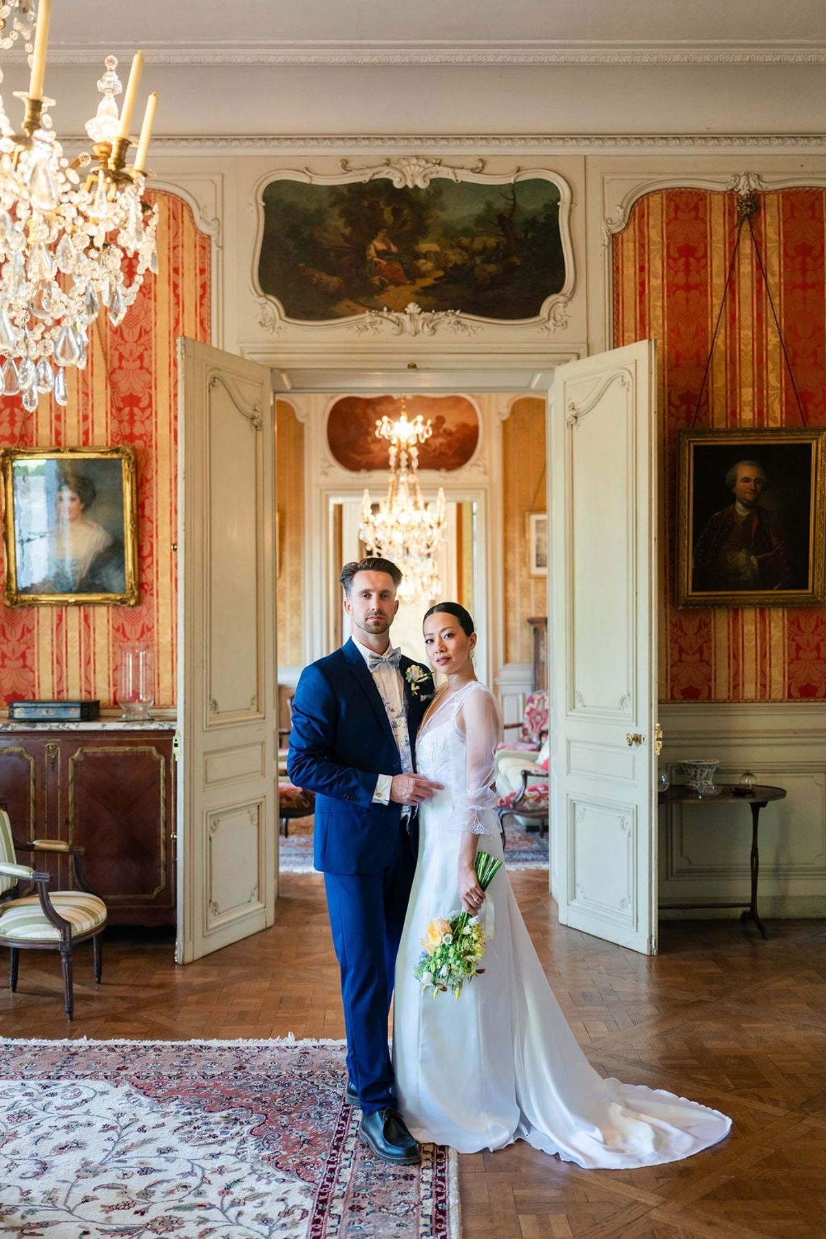 Bride and groom in the grand salon of Château du Terral with red silk walls, portraits and chandelier