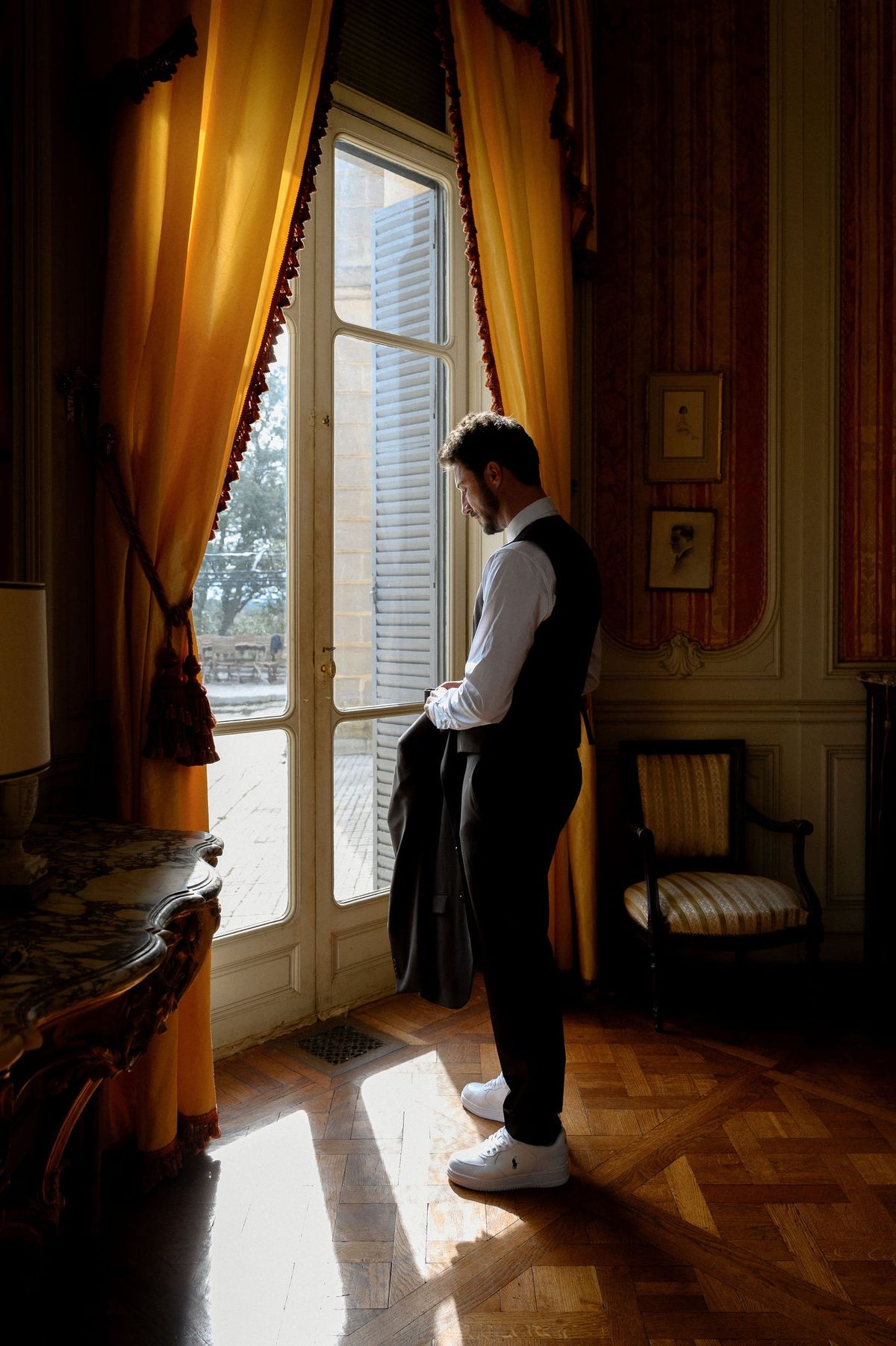 Groom holding his jacket by a tall window with gold curtains and parquet floor at Château du Terral