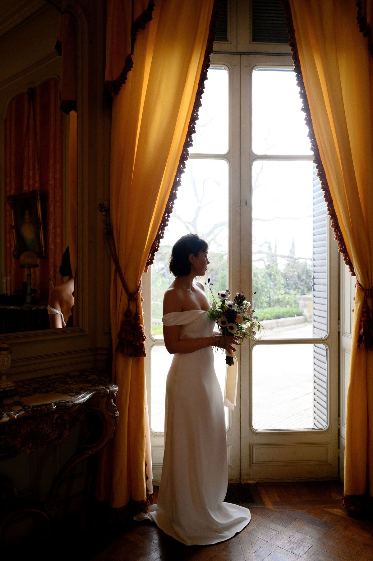 Bride with bouquet silhouetted at a tall window framed by gold curtains in Château du Terral's grand salon