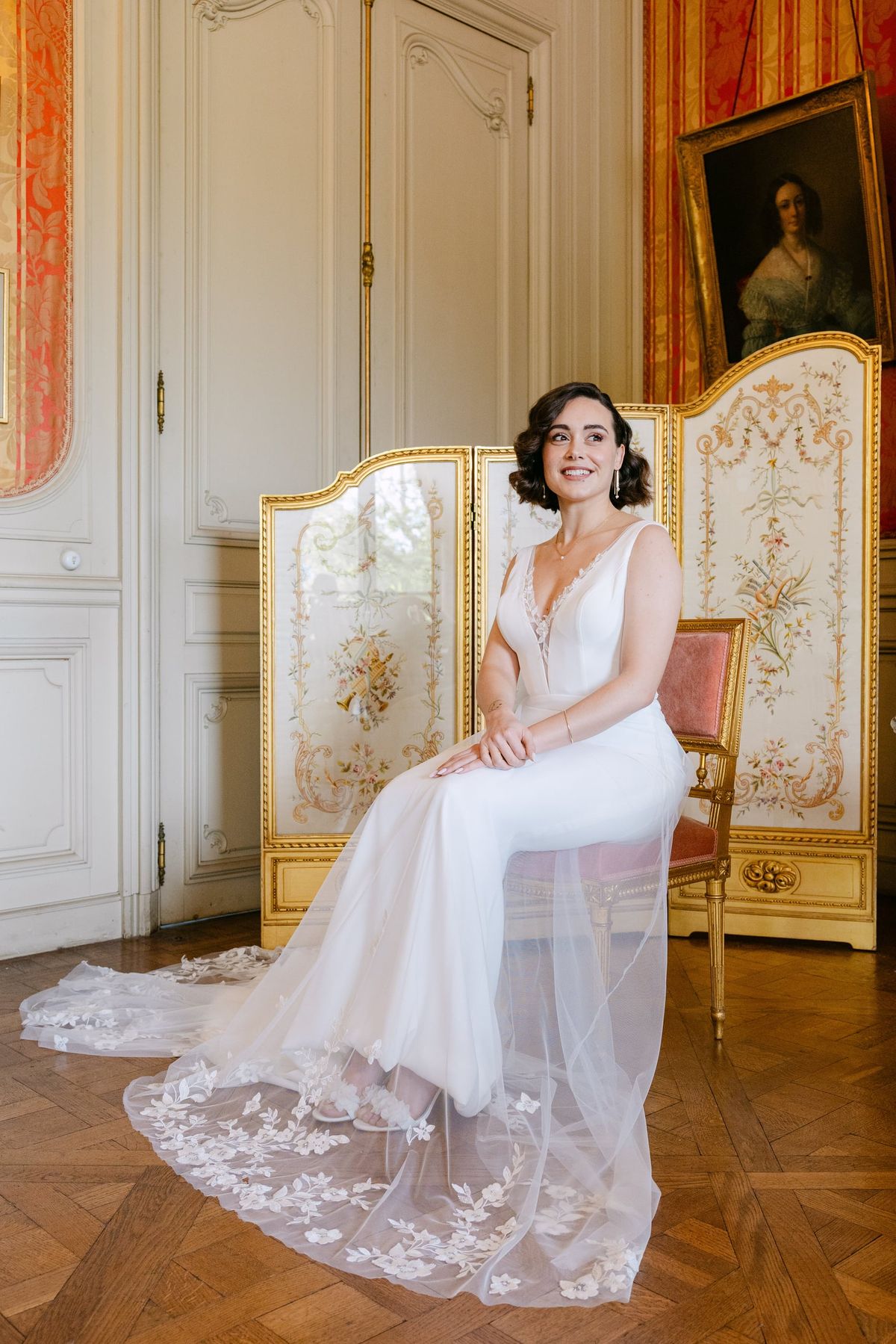 Seated bride in a lace-trimmed gown beside a painted folding screen and portrait at Château du Terral