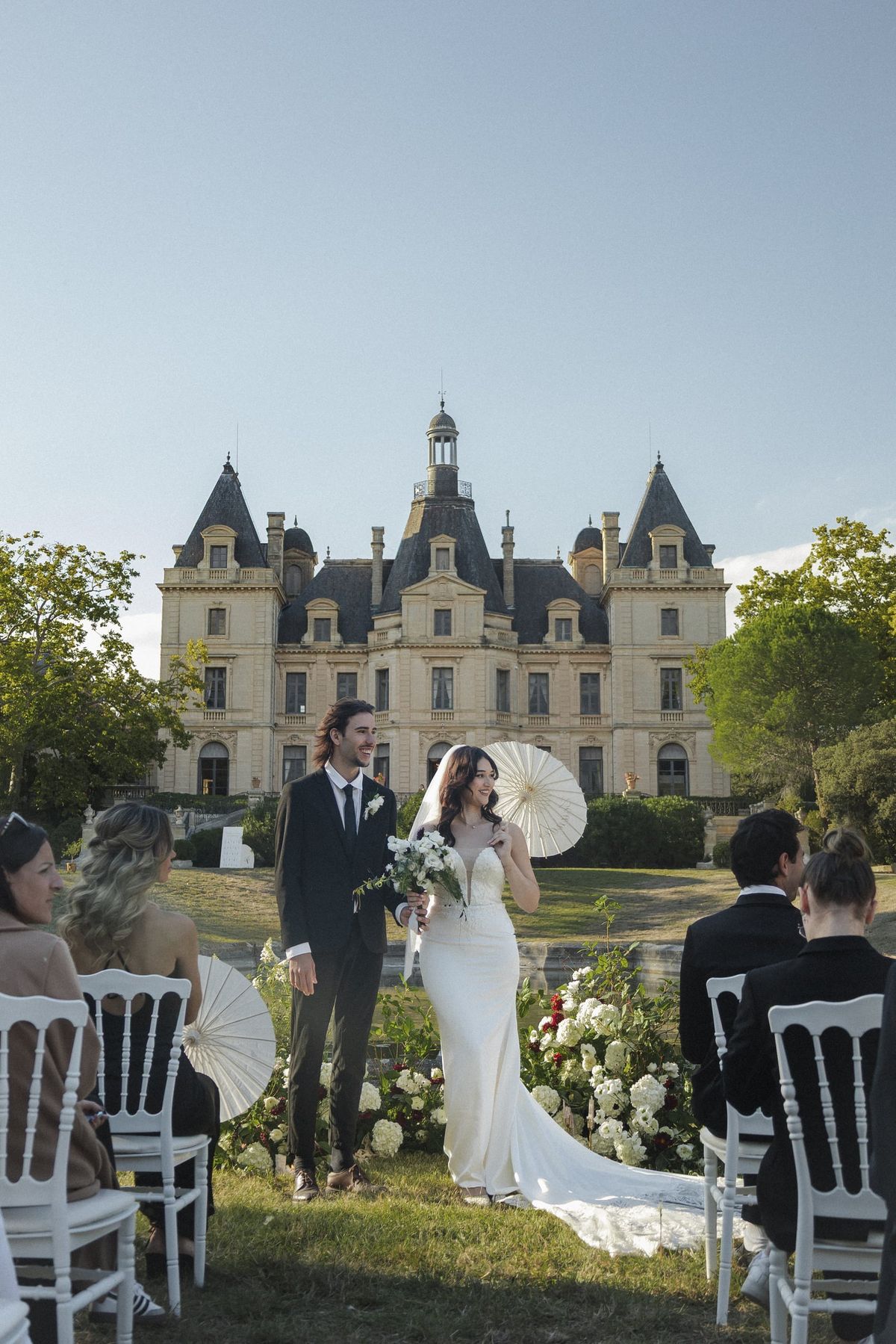 Outdoor wedding ceremony on the lawn before Château du Terral, guests holding parasols