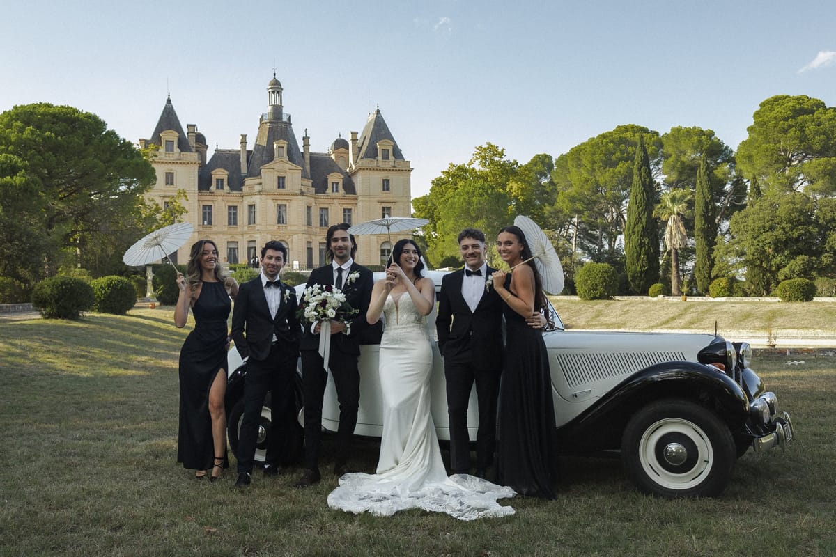 Wedding party with parasols beside a vintage car on the lawn before Château du Terral