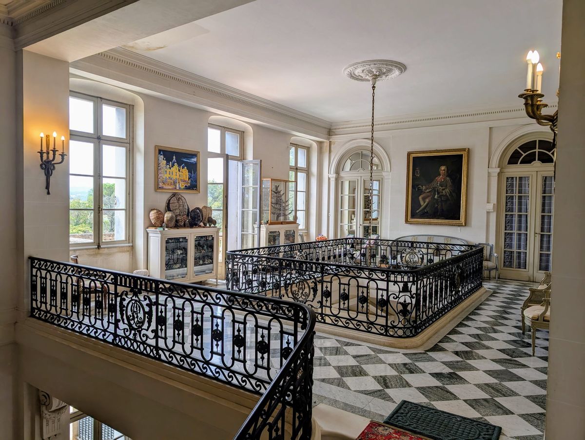 Sunlit upstairs landing at Château du Terral with iron balustrades, tall windows and chequered marble floor