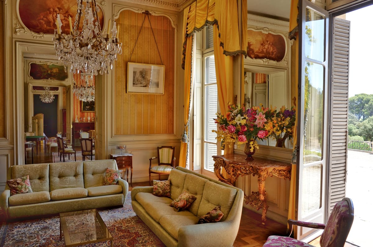 Sunlit salon at Château du Terral with painted ceiling panels and a floral display by open windows