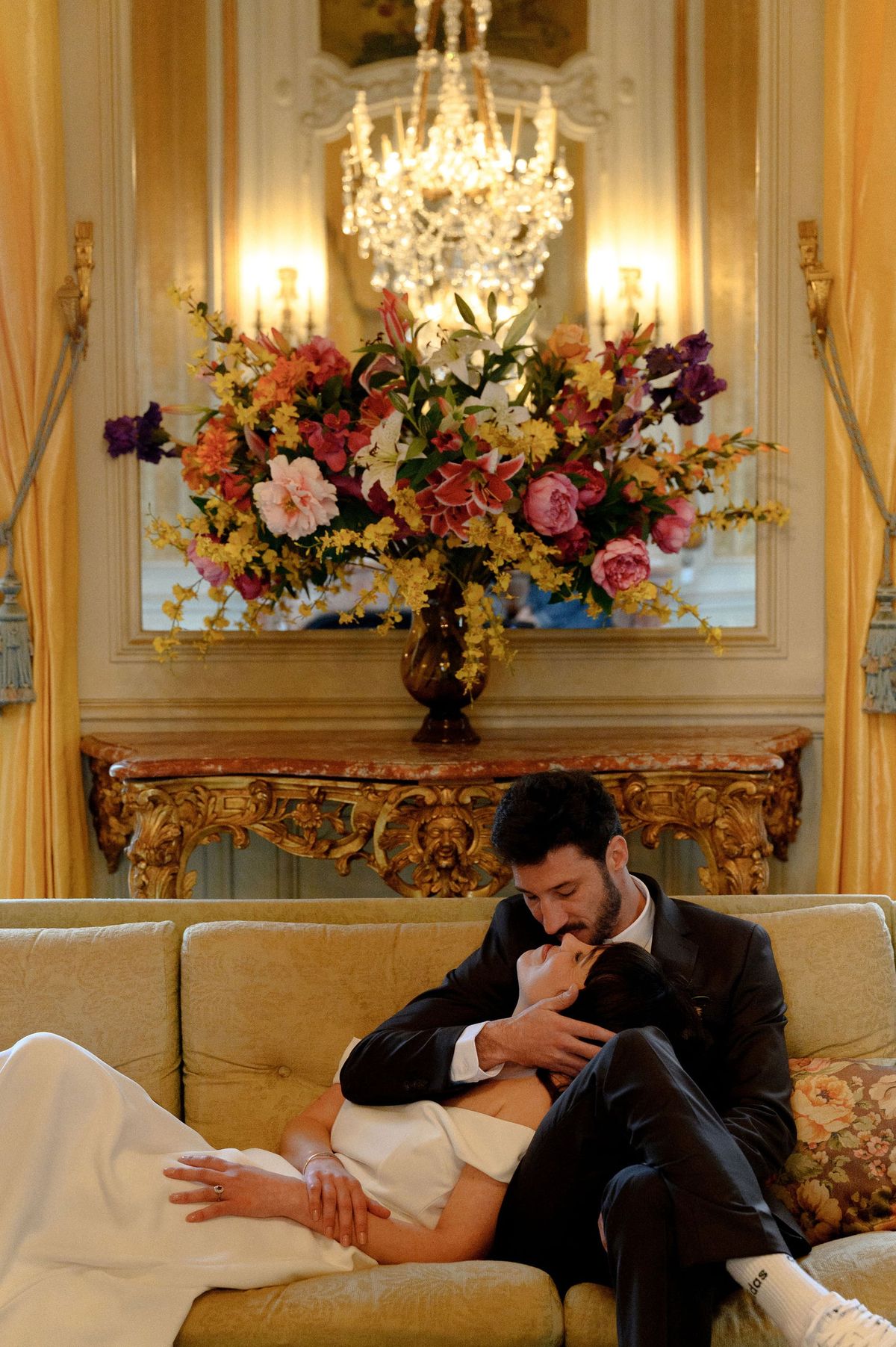 Couple embracing on a sofa beneath a floral display and mirror in a Château du Terral salon