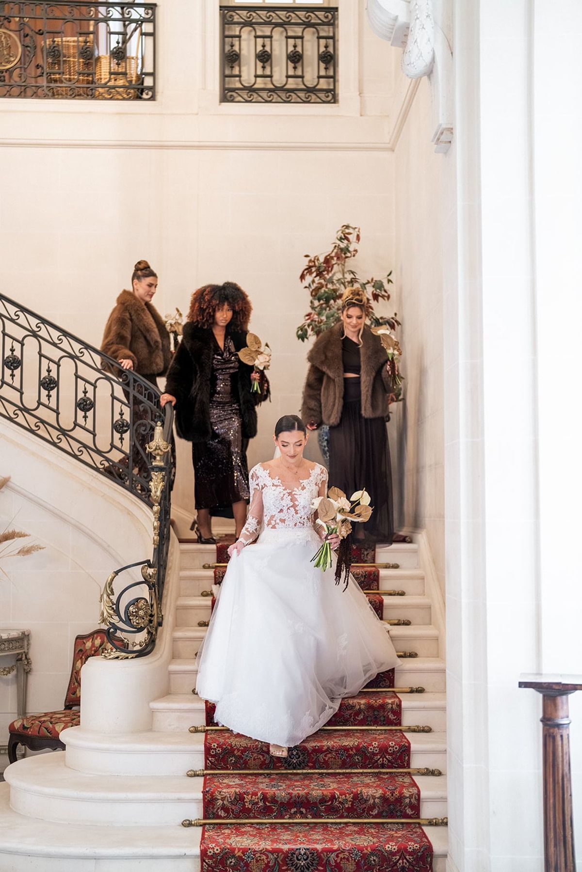 Bride descends the red-carpeted staircase of Château du Terral with friends in fur coats behind