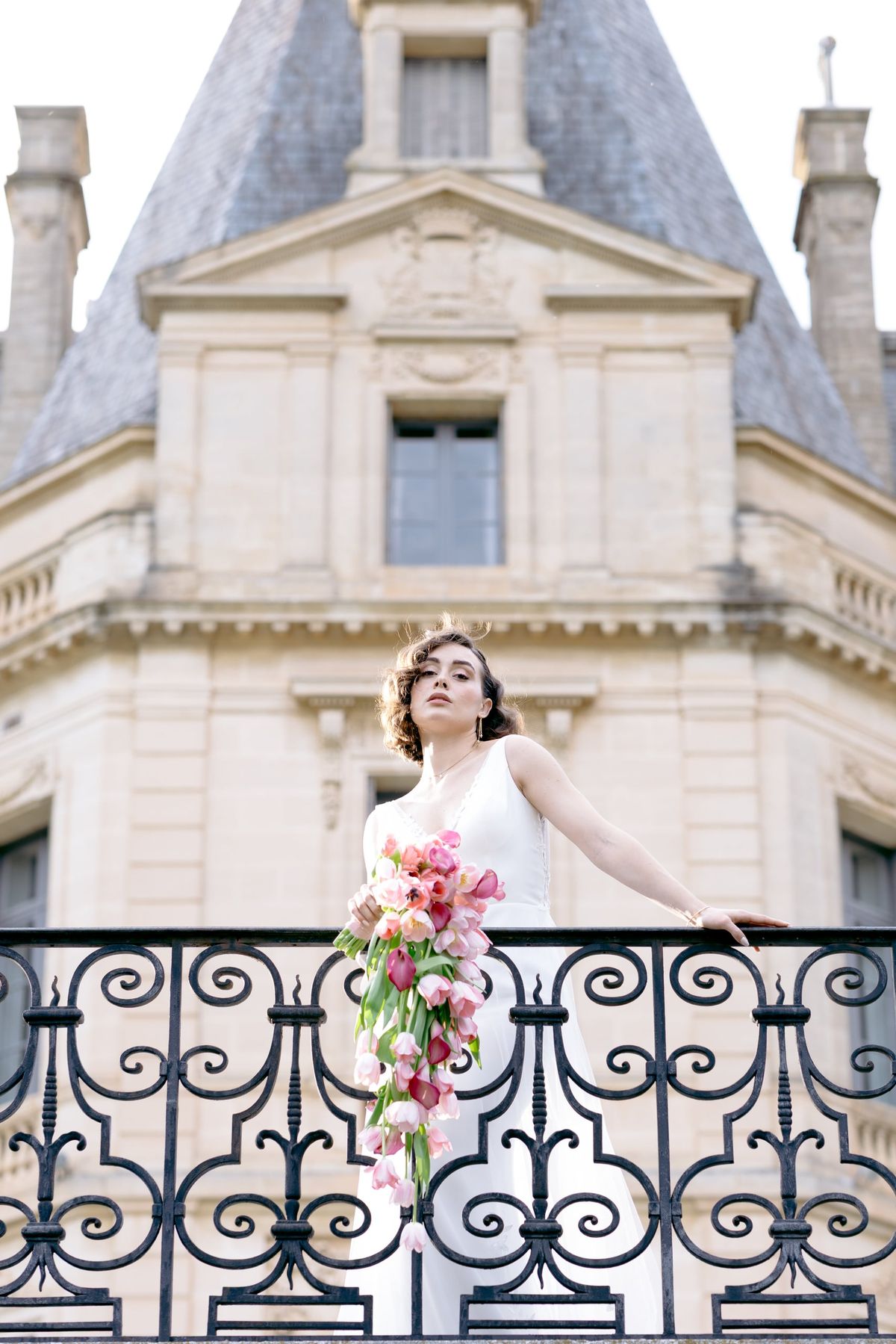 Bride with a cascading pink bouquet on an iron balcony below a Château du Terral turret