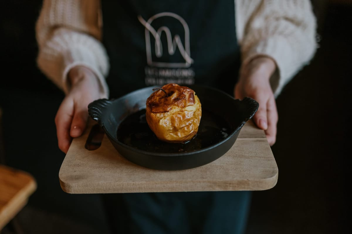 Staff member in a Les Maisons du Bonheur apron serving a baked apple dessert on a wooden board