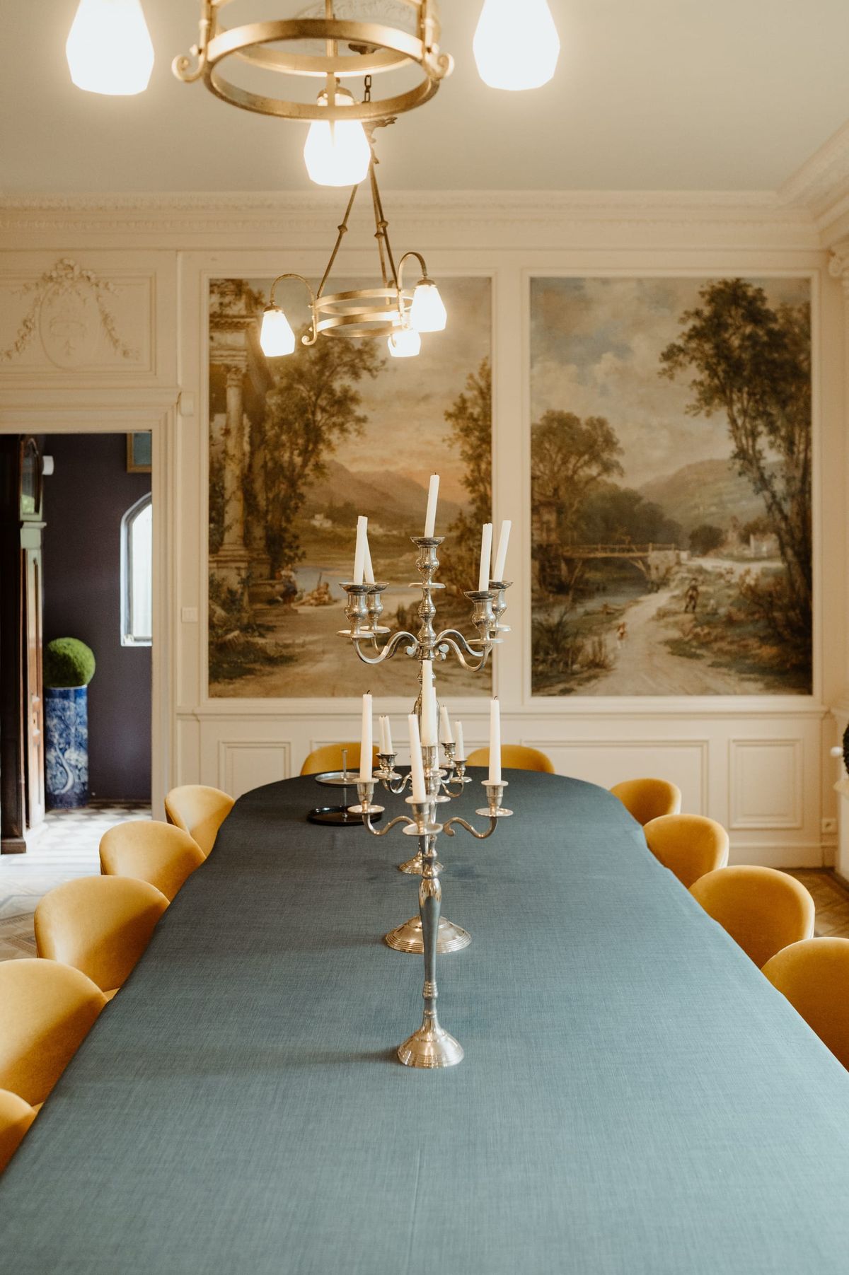 Long dining table with a silver candelabra beneath framed landscape murals and mustard velvet chairs
