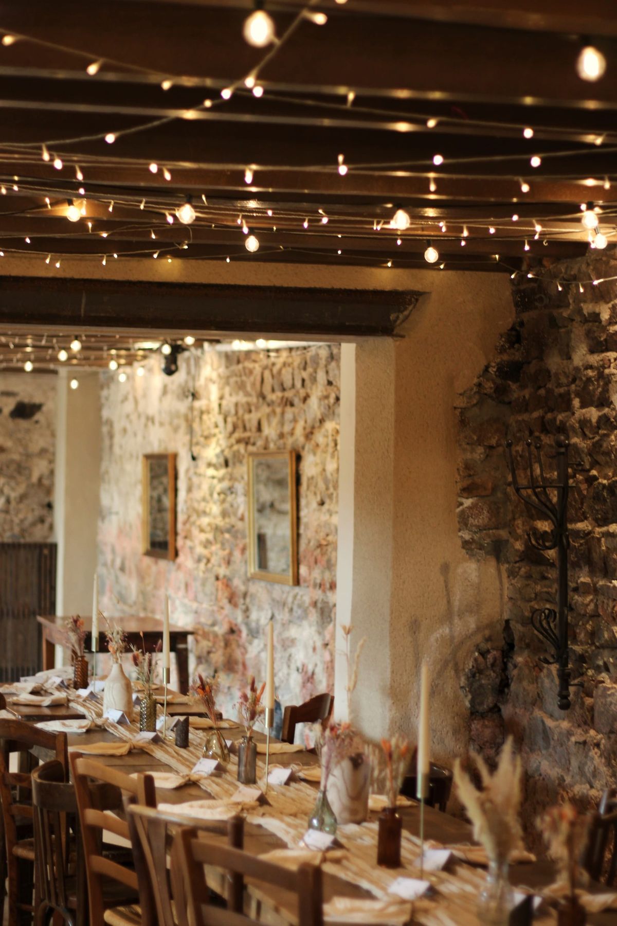 Stone-walled reception room strung with festoon lights above tables set with dried flower centrepieces