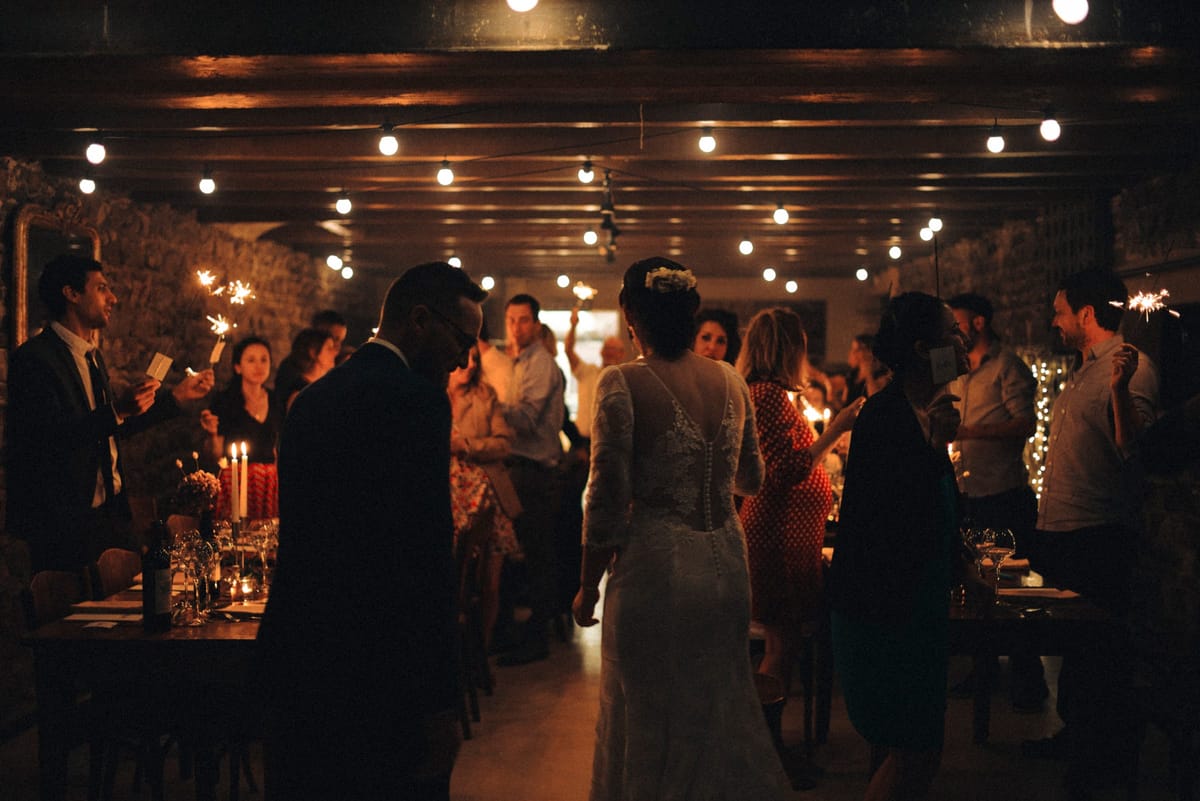 Evening reception with guests holding sparklers under festoon lights in the stone-walled hall