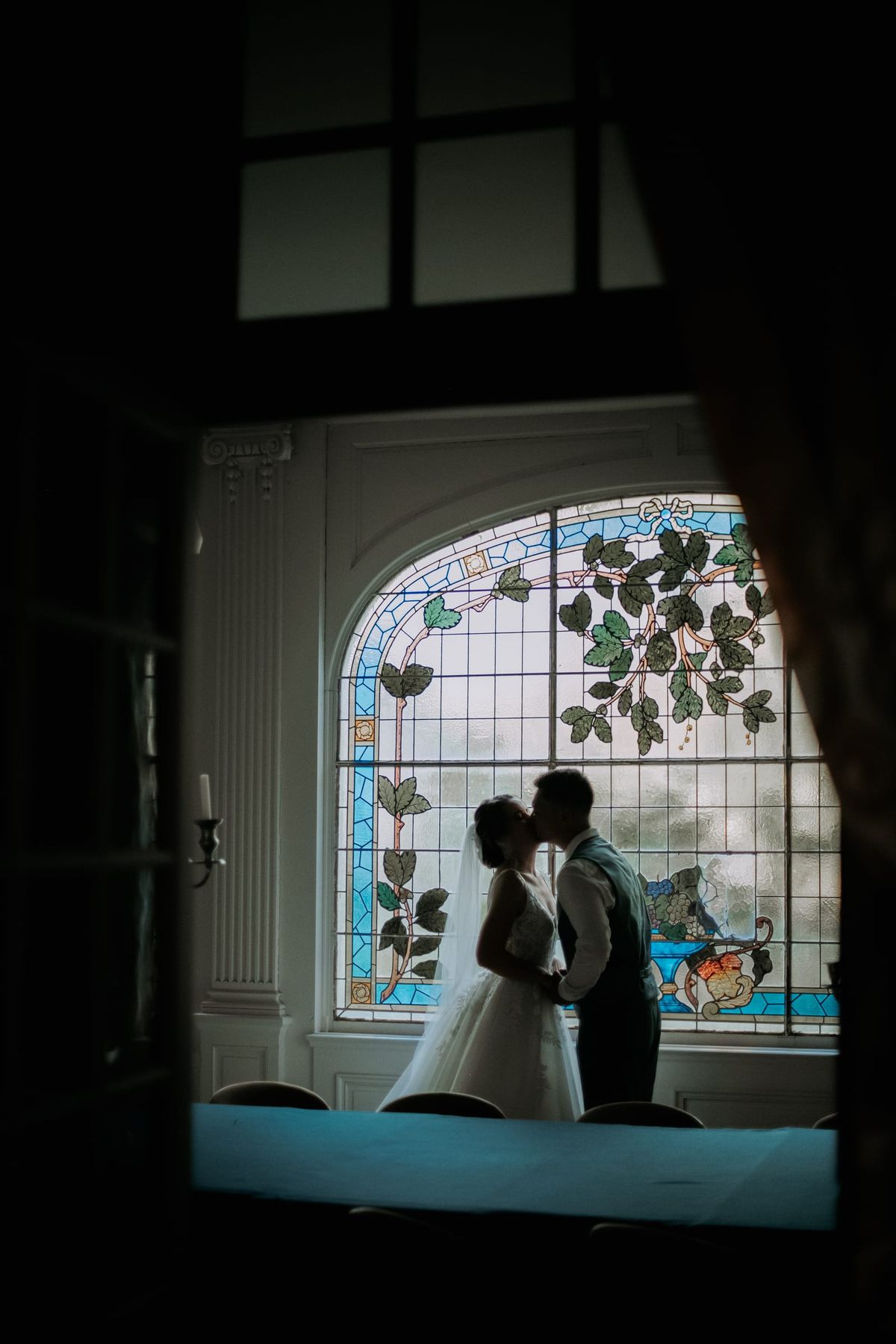 Silhouette of bride and groom kissing in front of a stained-glass window, framed by a doorway