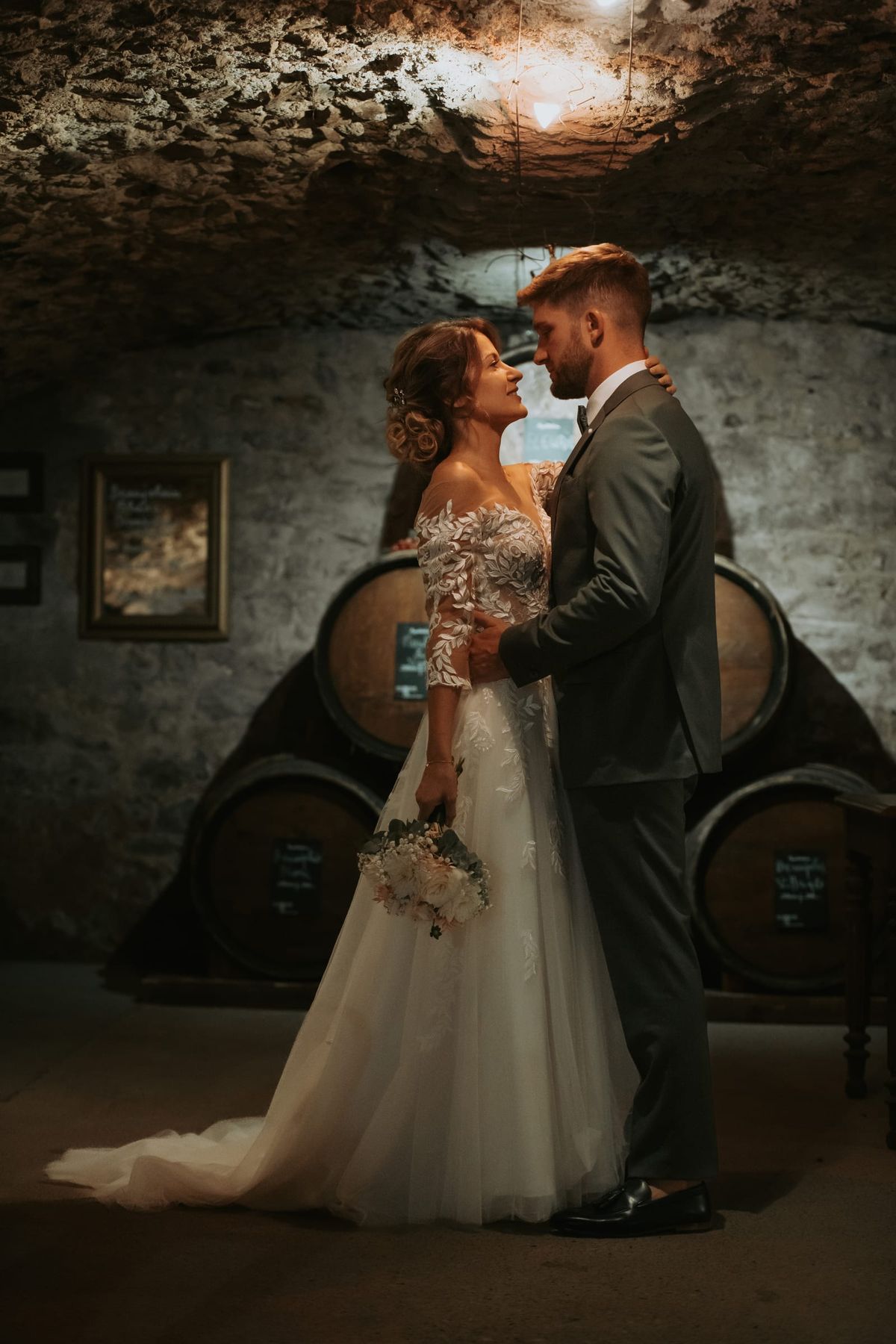 Bride and groom embracing inside the stone wine cellar among oak barrels