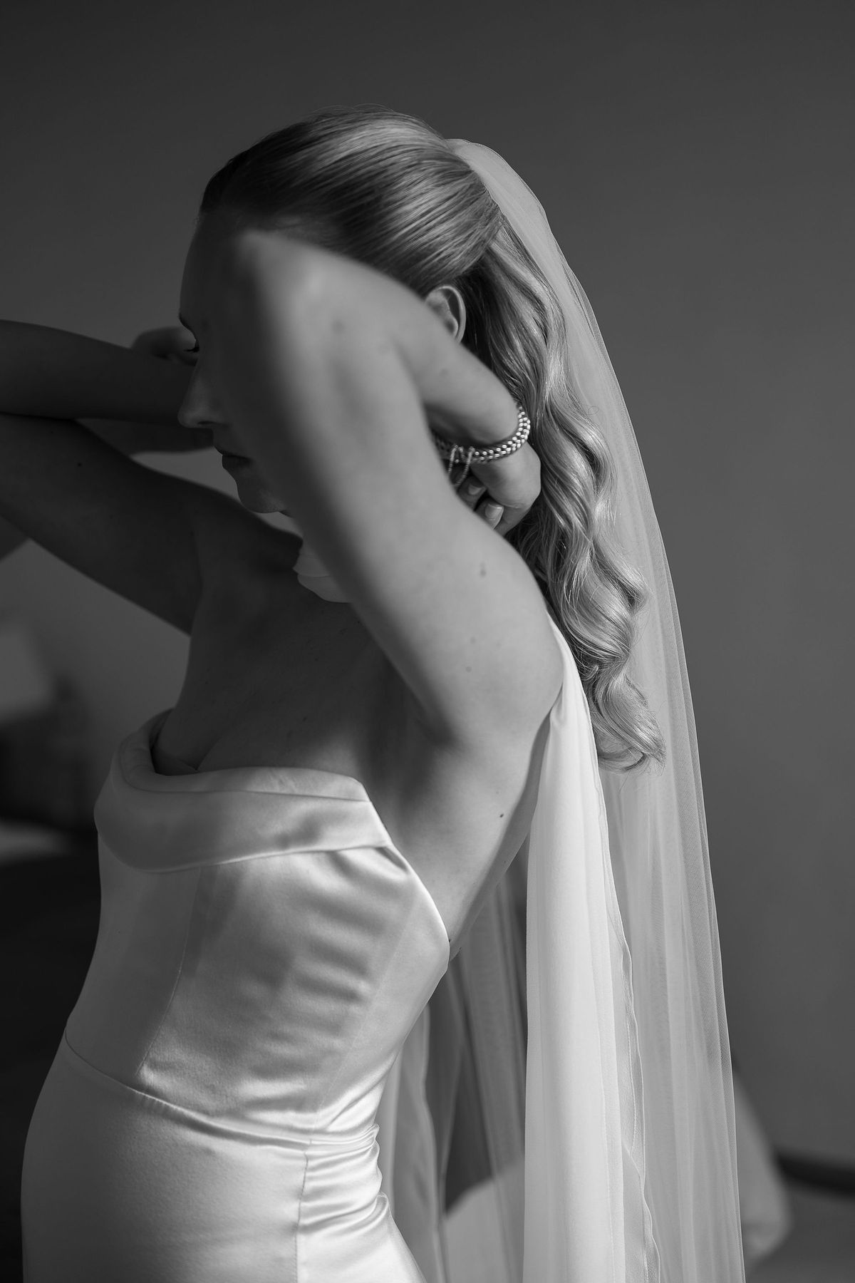 Black and white portrait of Louise adjusting her hair, wearing a silk bustier gown and a full length veil