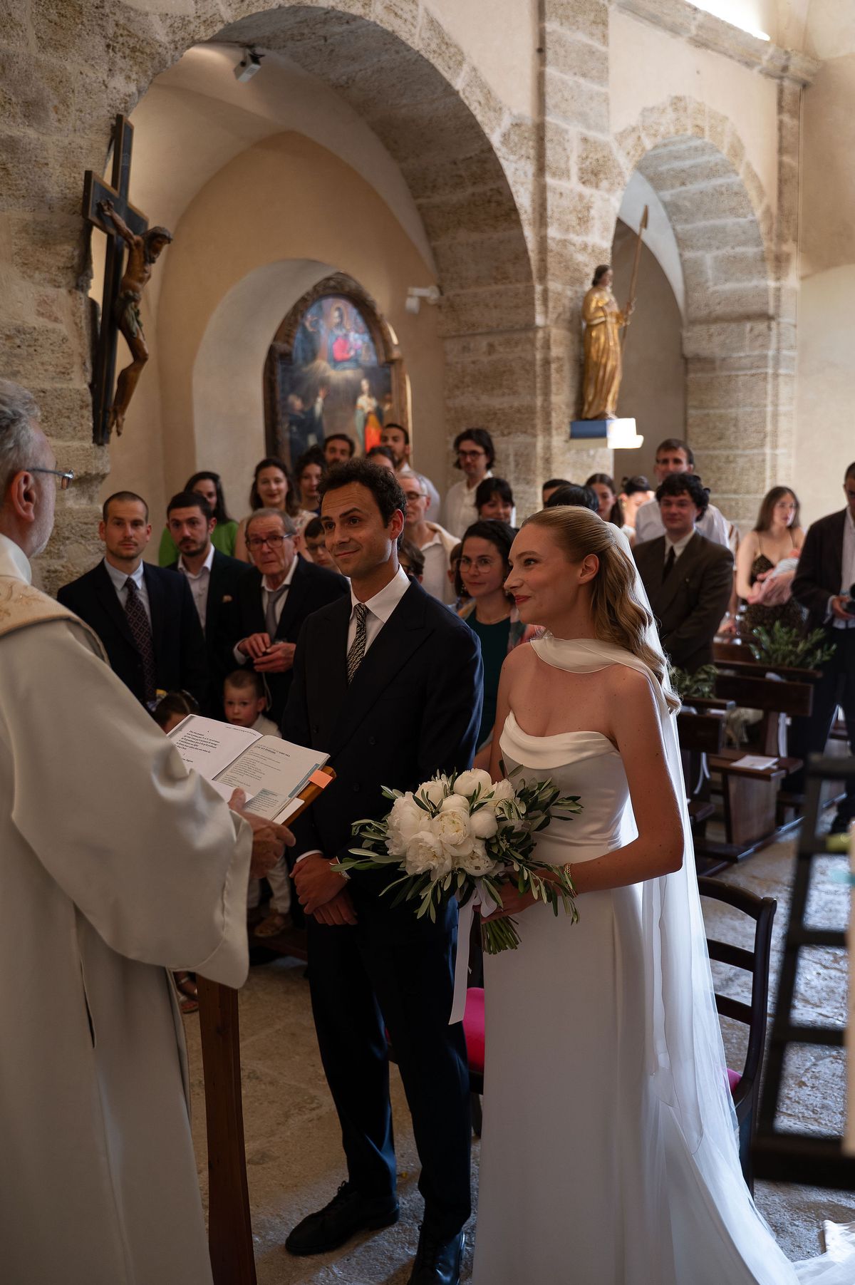 Louise and Lucas standing before the priest during their religious ceremony in a stone village church