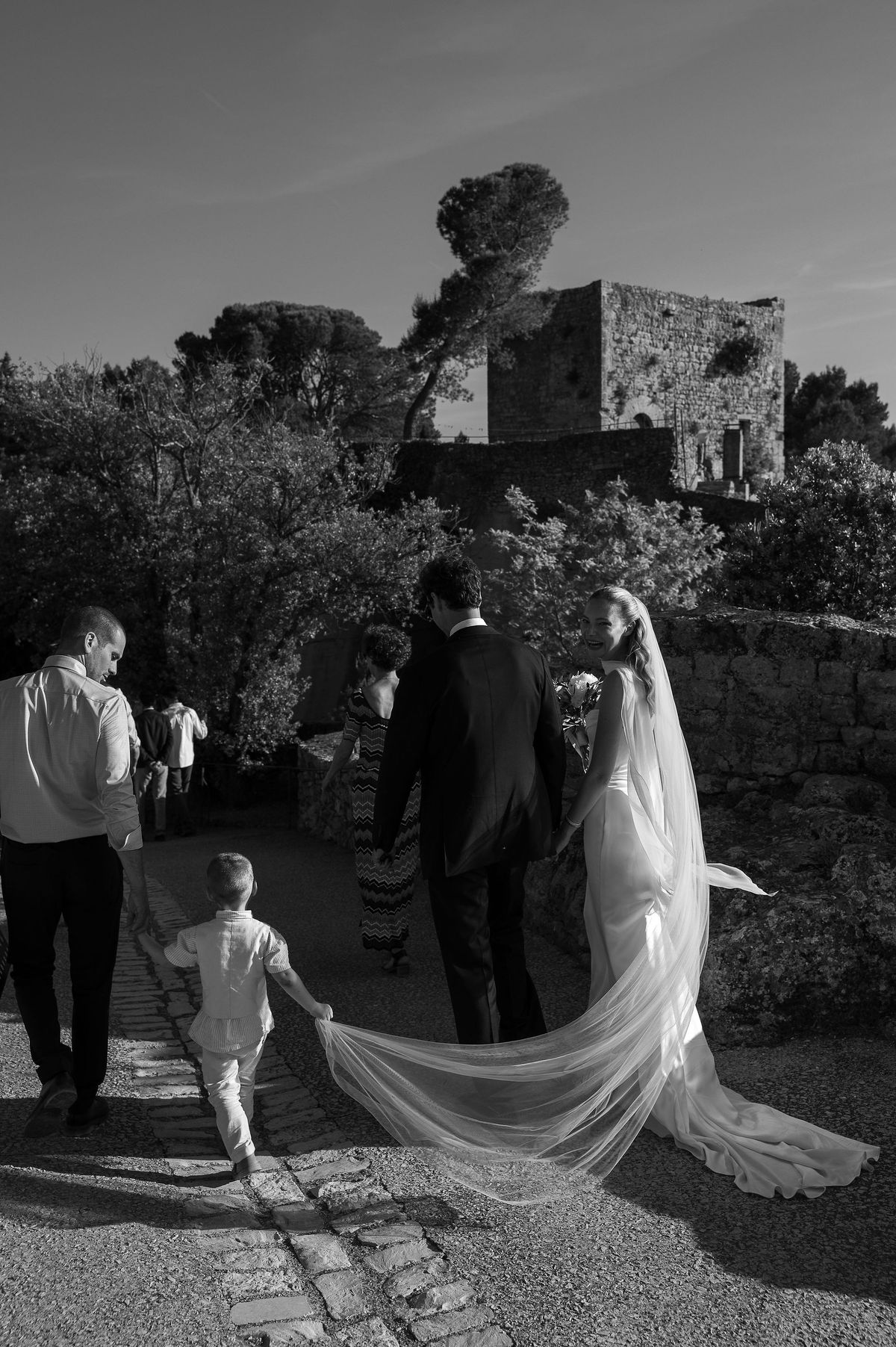 Black and white photograph of a child carrying Louise's veil as the couple walk through the village