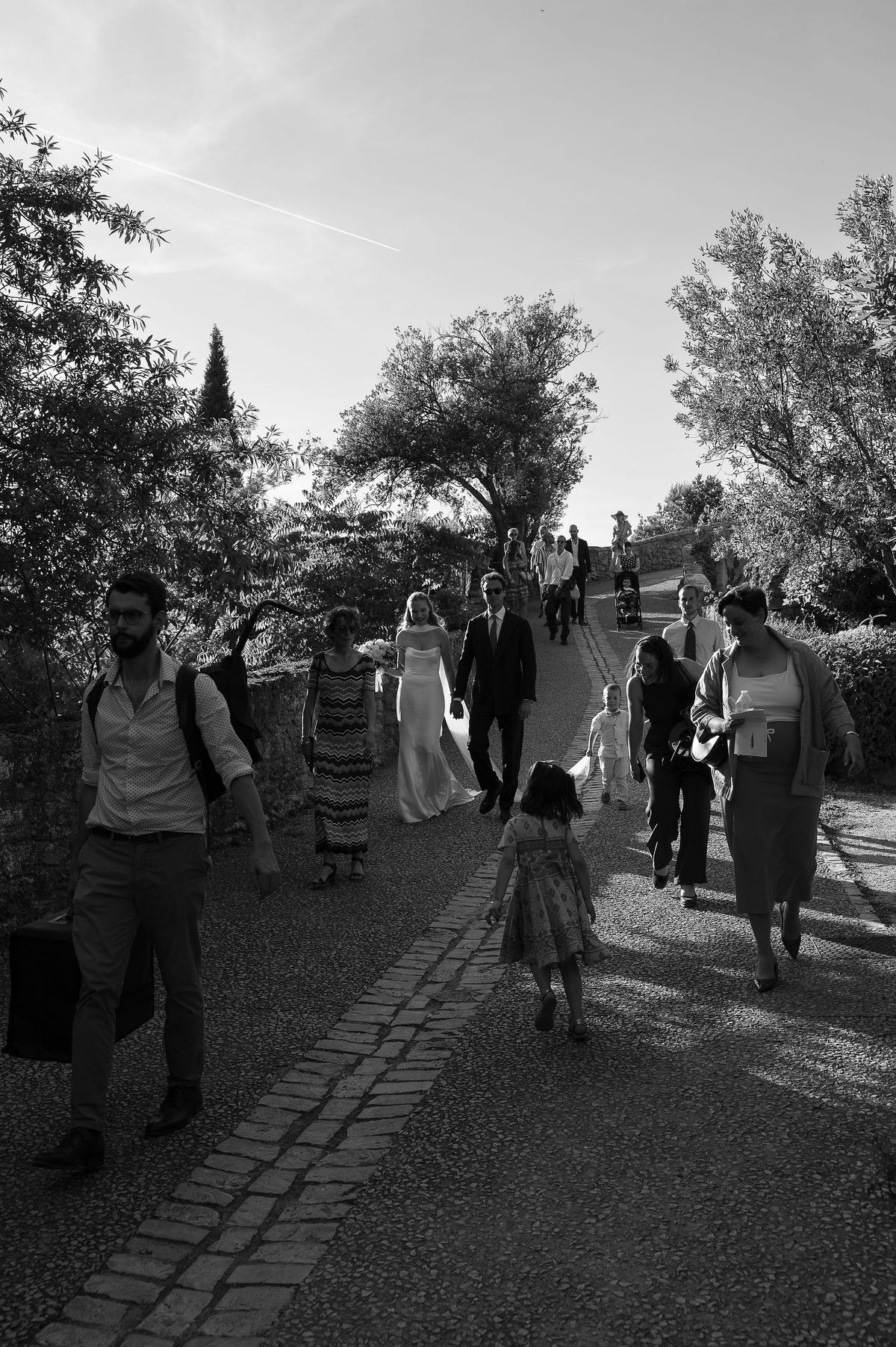 Black and white photograph of guests walking down the village lane beneath olive trees