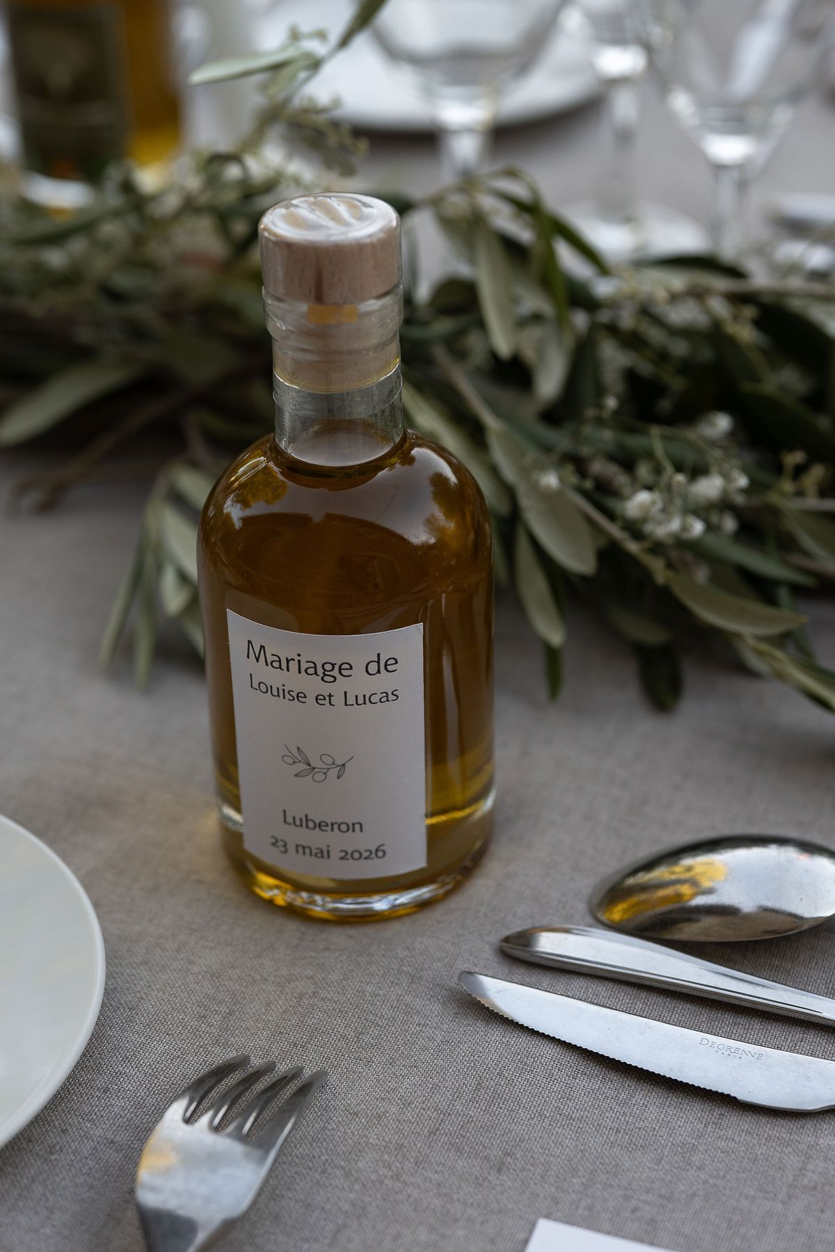 A bottle of olive oil with a printed wedding label, set among olive branches on the table