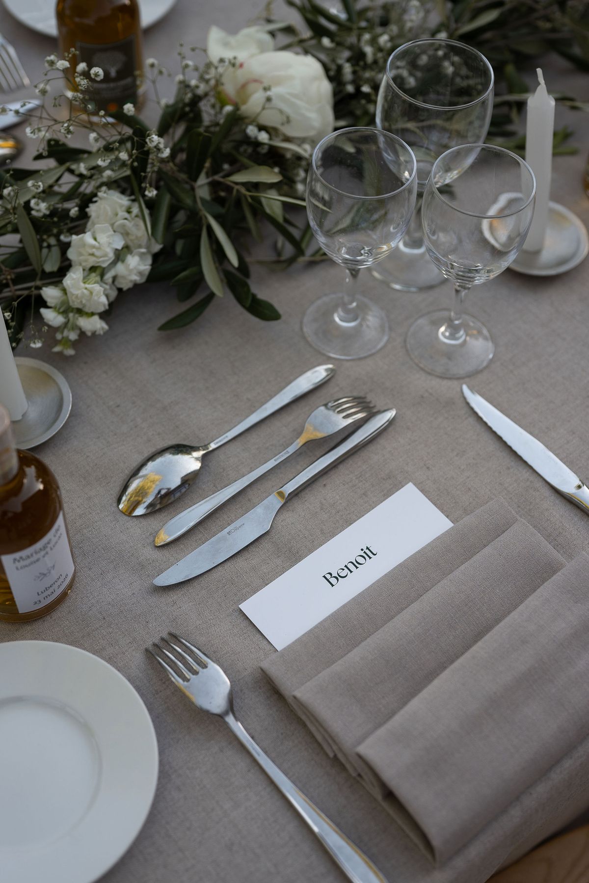 Place setting with a printed name card, olive branches and white flowers on a linen tablecloth