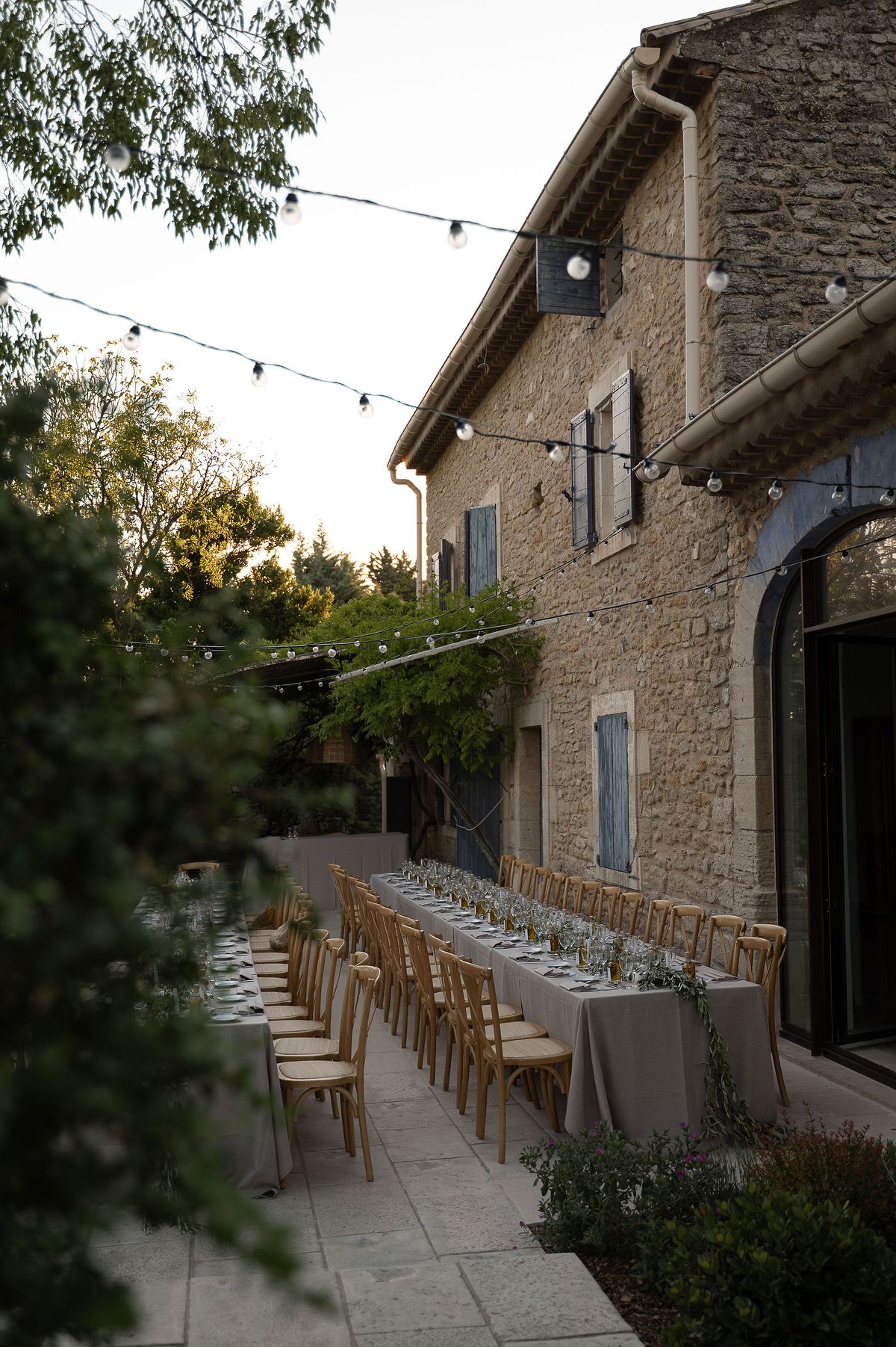 The long candlelit table beneath string lights beside the stone house at dusk