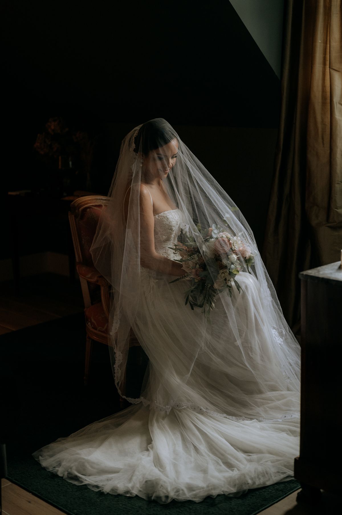 Bride getting ready, seated with her veil and bouquet in a dimly lit interior