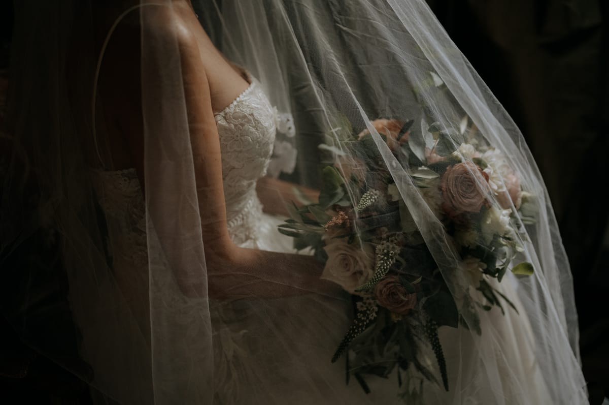 Close-up of a bride beneath her veil, holding a rose and eucalyptus bouquet