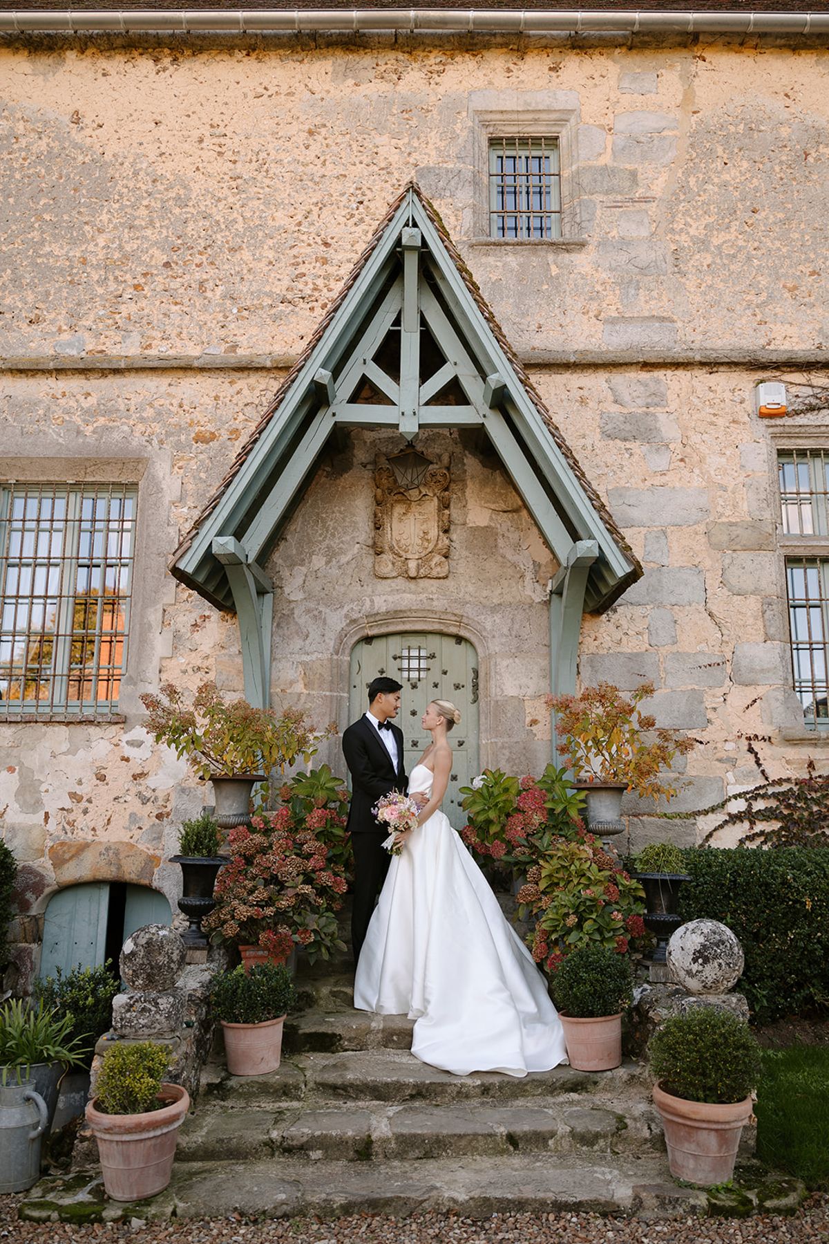 Couple portrait beneath the manor's timber-framed entrance porch, flanked by potted hydrangeas
