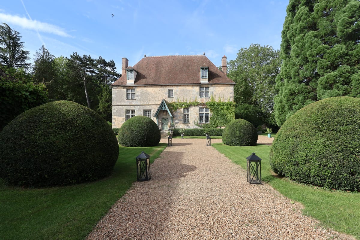 Wide exterior shot of the manor's facade framed by clipped topiary spheres along the gravel drive