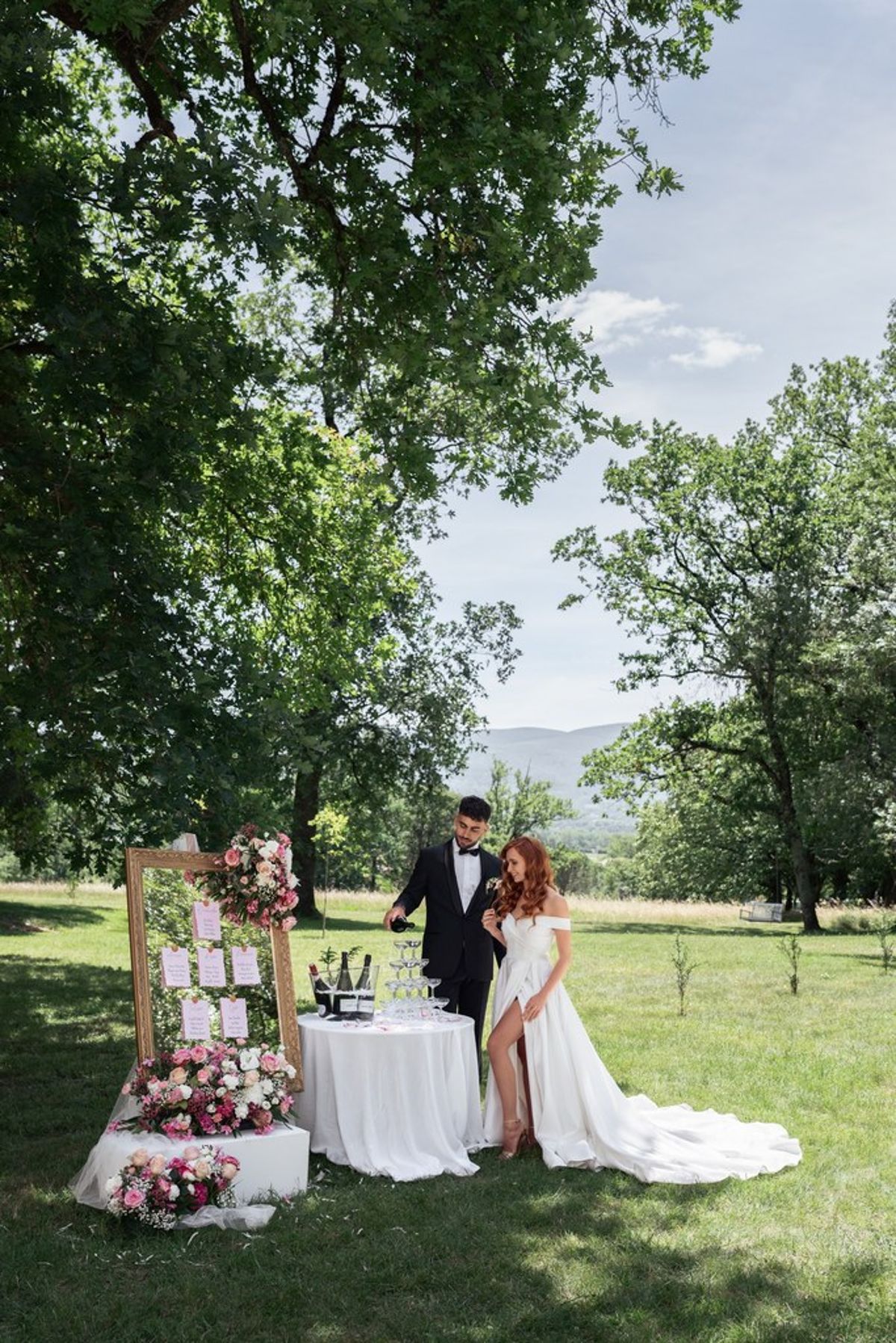 Blush and fuchsia styled shoot at Château du Causse, Occitanie, the couple beside a gilt frame of table cards and a flower table in the park