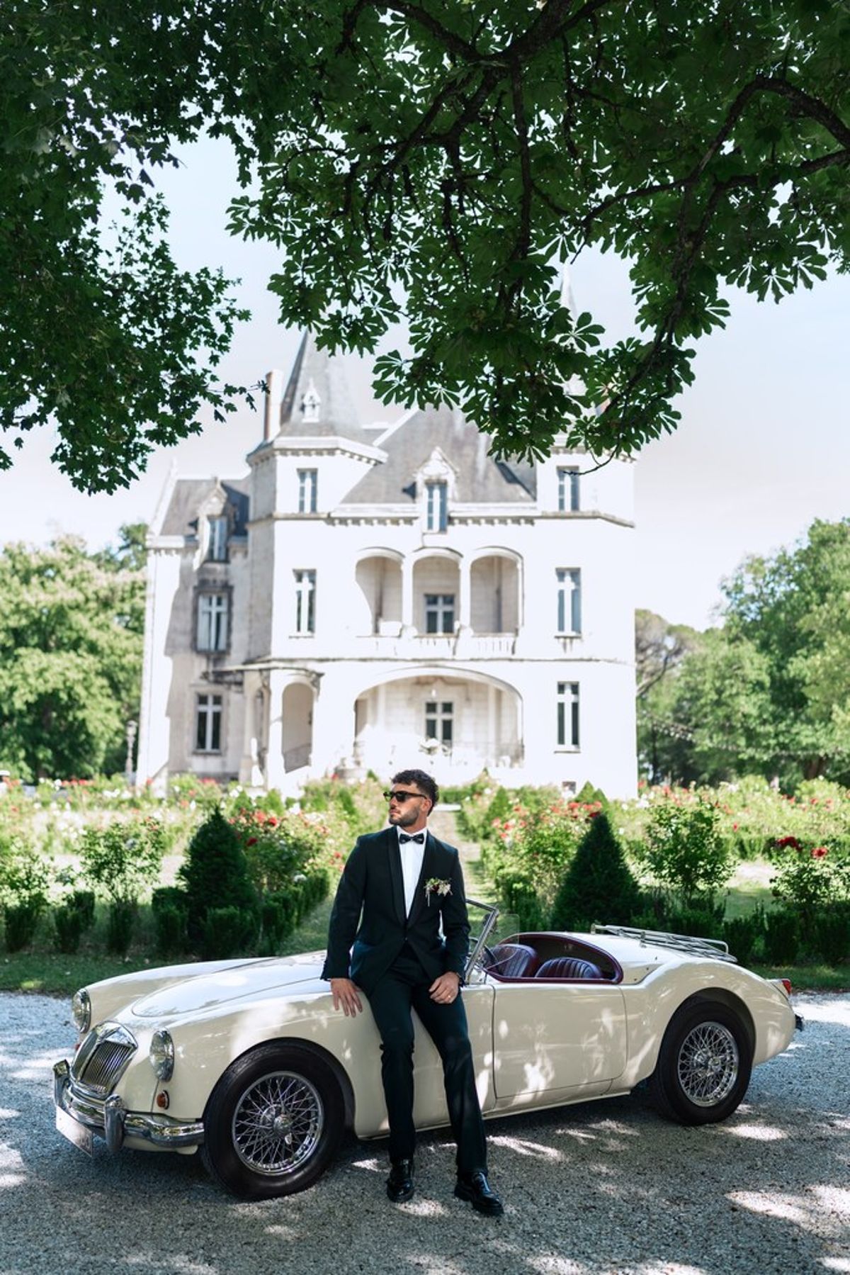 Blush and fuchsia styled shoot at Château du Causse, Occitanie, the groom beside a vintage car on the drive