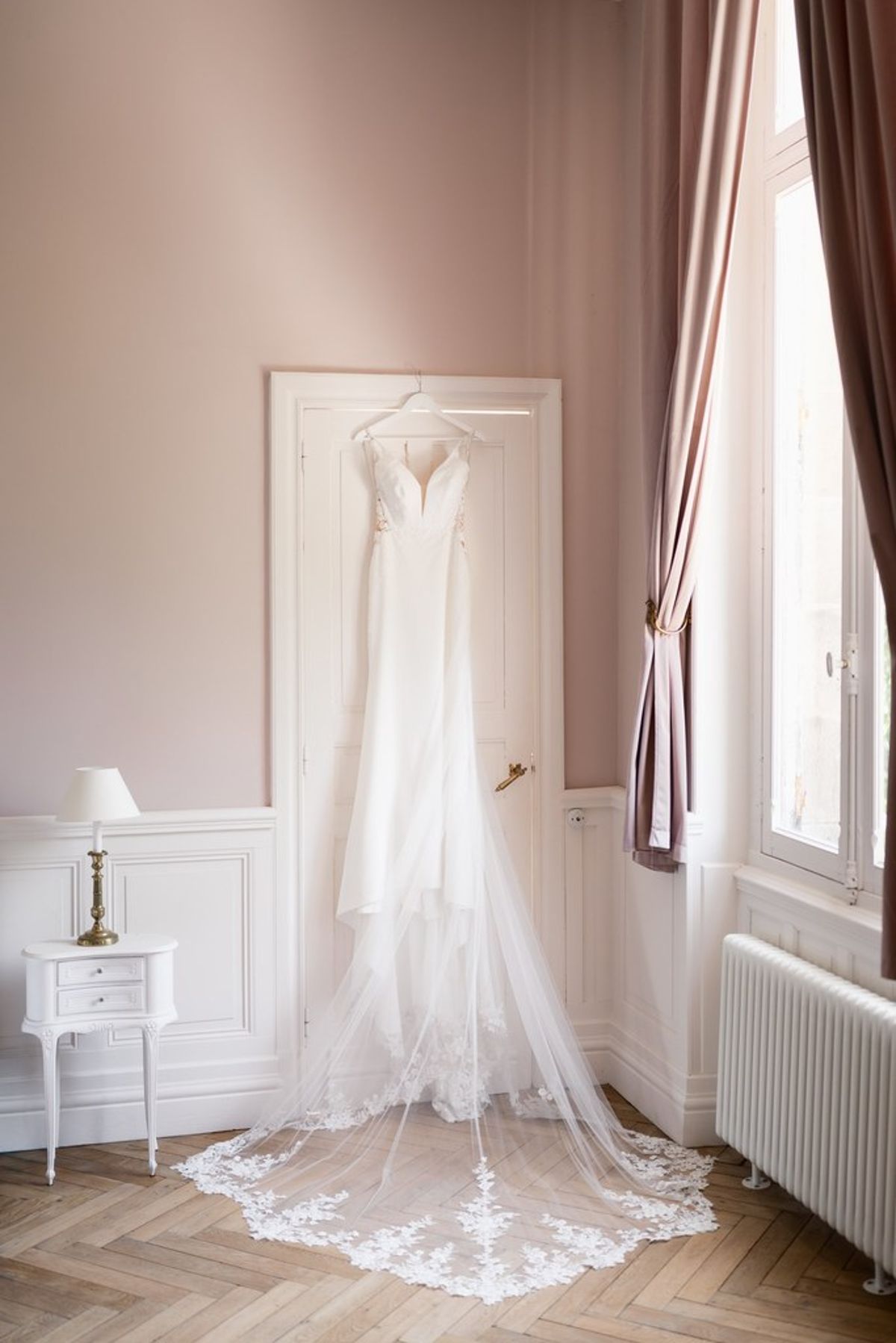 Blush and fuchsia styled shoot at Château du Causse, Occitanie, the wedding gown hanging by a tall window in a pale pink bedroom