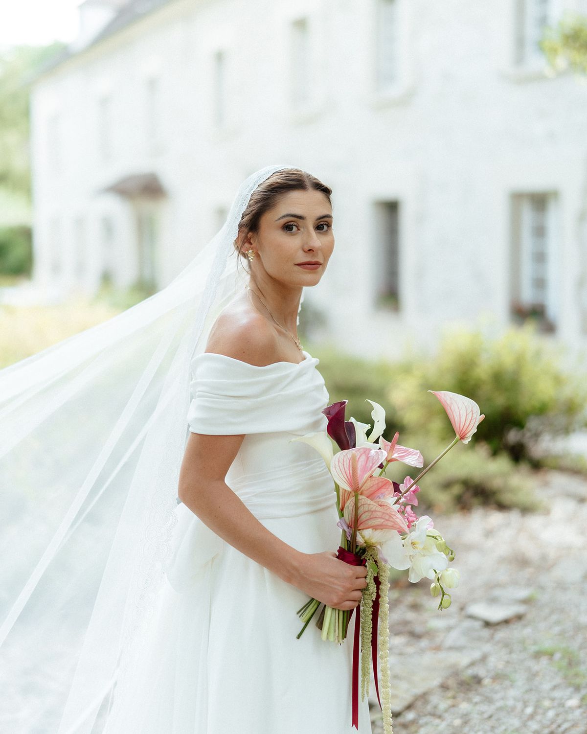 Bride in an off the shoulder draped gown and long veil holding a pink anthurium bouquet
